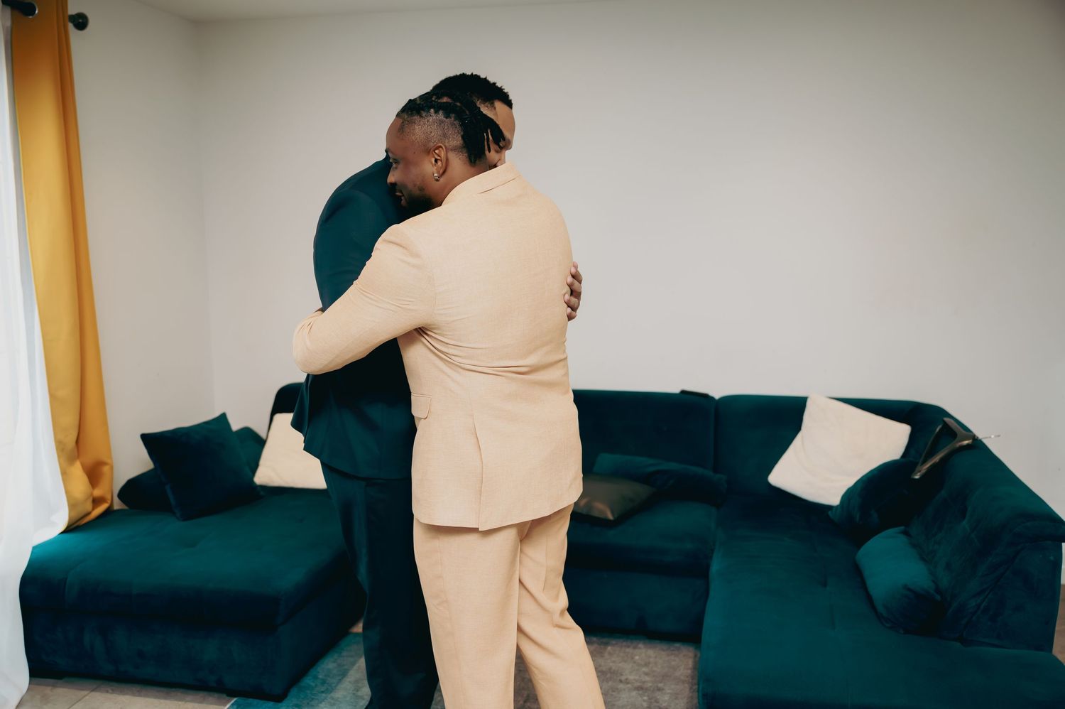 Two men in formal suits sharing a warm embrace in a living room before a wedding.