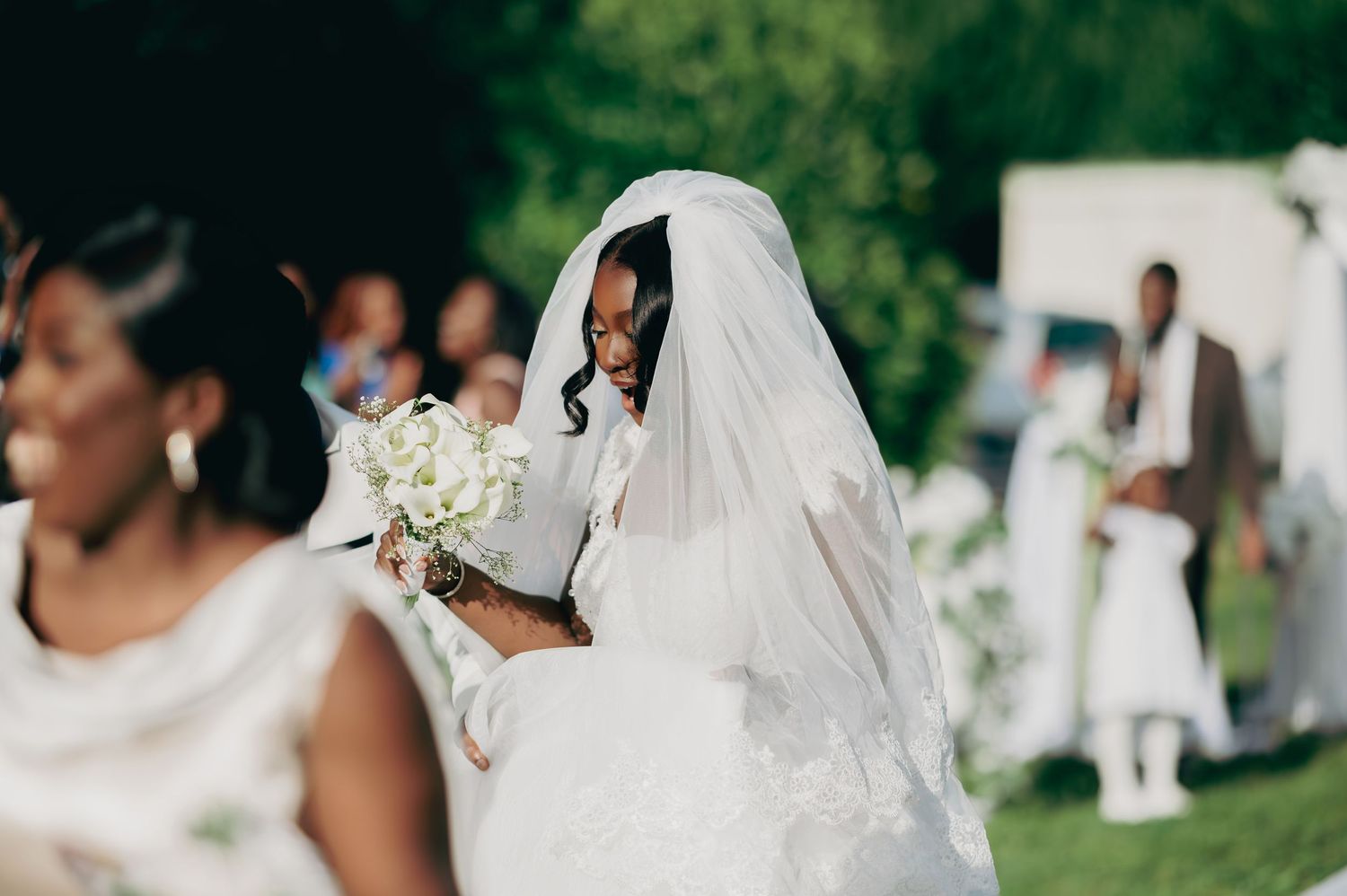 Bride in white wedding gown and veil holds bouquet of white flowers at outdoor ceremony.