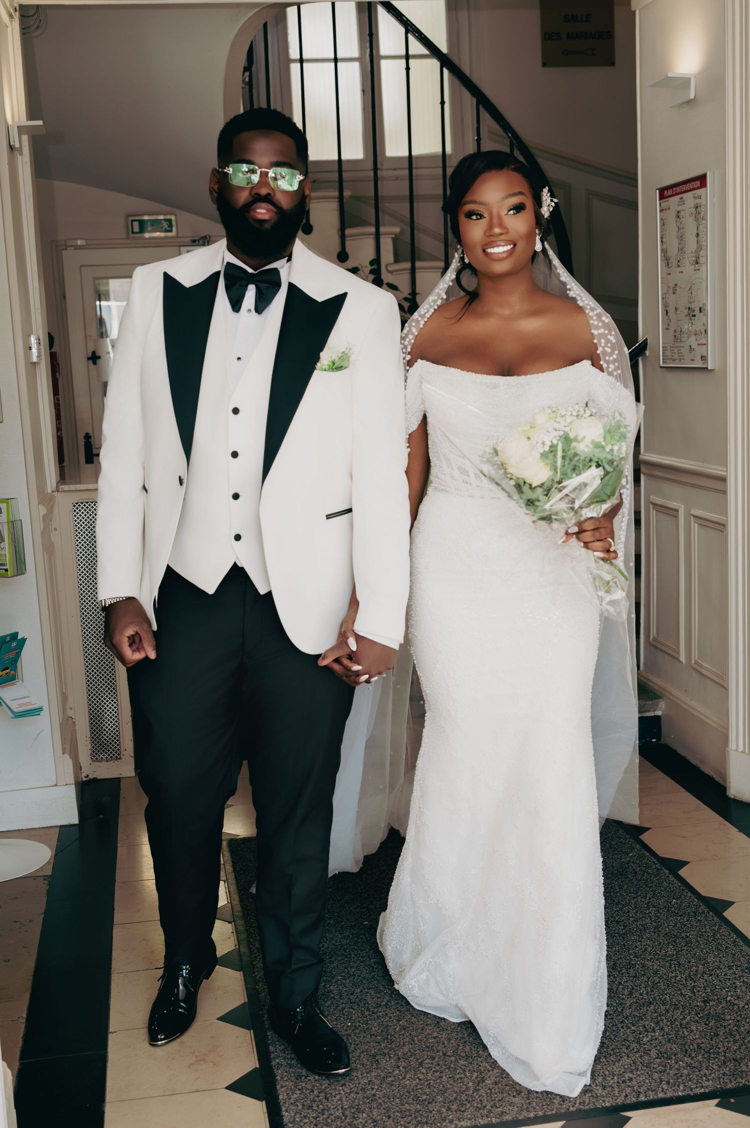 Stylish groom in white tuxedo with sunglasses and bride in off-shoulder lace gown pose holding hands.