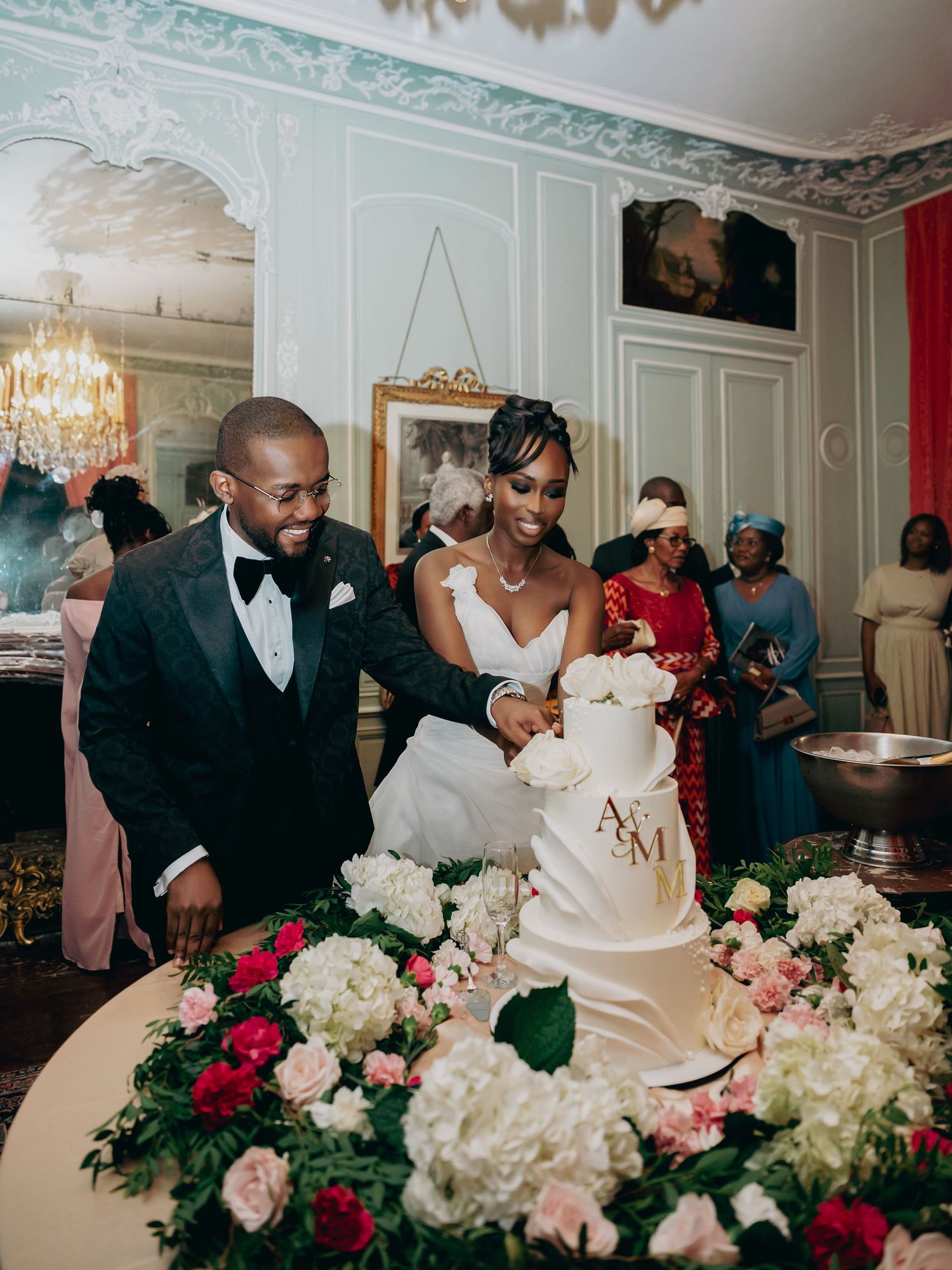 Bride and groom cutting a white floral wedding cake adorned with gold monogram initials at an elegant reception.