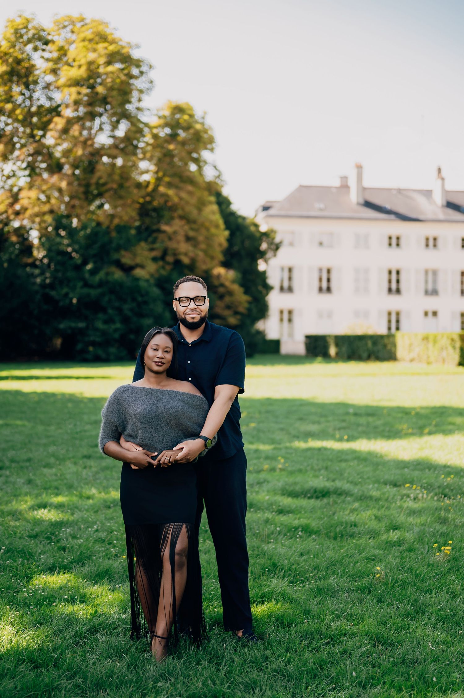 Couple embraces on green lawn with grand white estate building in the background.