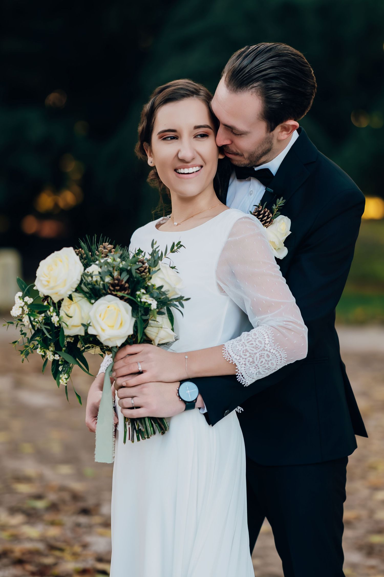 Groom tenderly embraces smiling bride holding white rose and greenery bouquet in romantic outdoor portrait.
