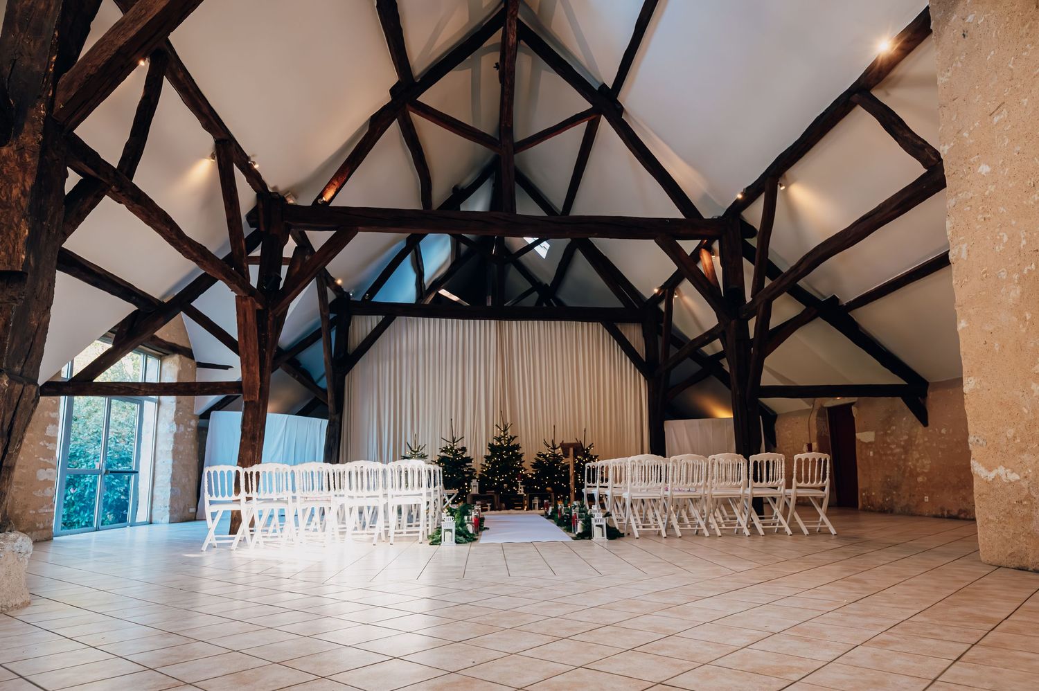 Elegant barn wedding venue with white chairs, exposed dark wood beams, and small Christmas tree altar decoration.