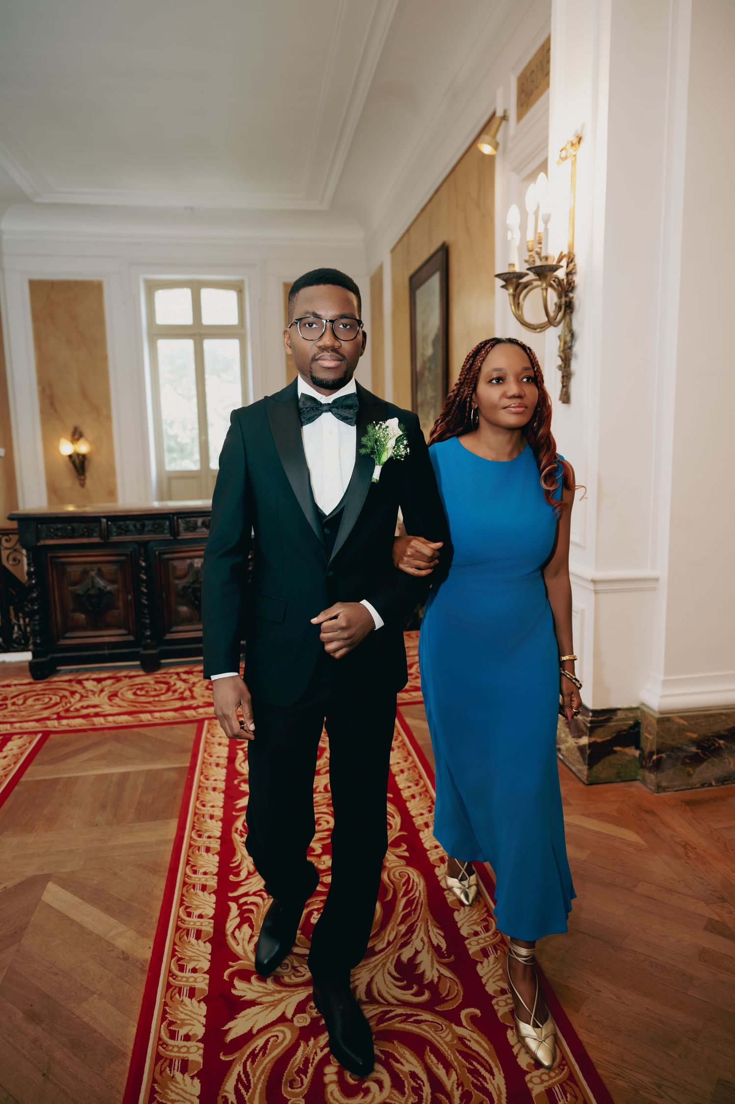 Groom in dark green tuxedo and bride in blue dress walk through ornate wedding venue hallway.