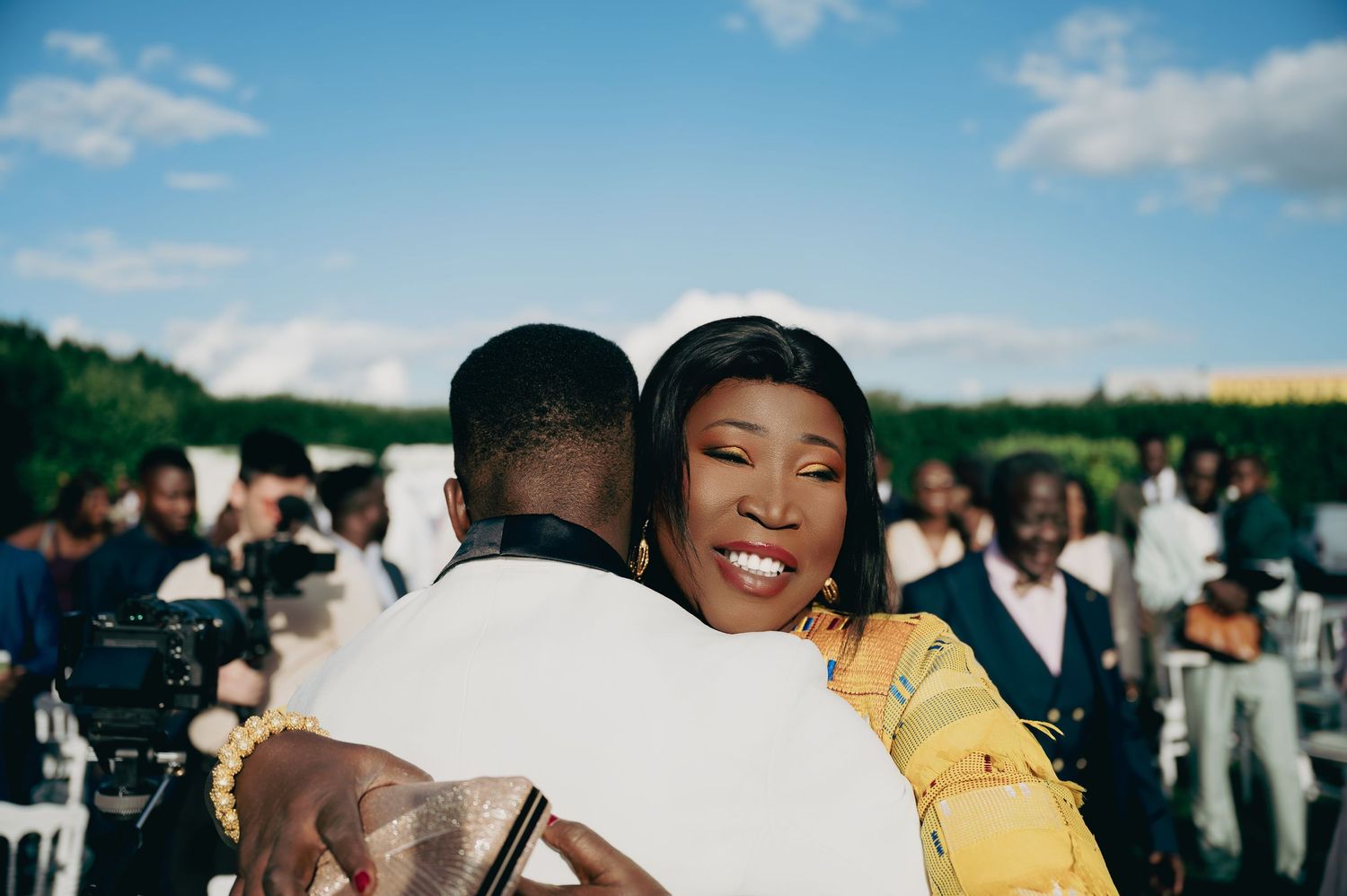 Woman in yellow dress shares warm embrace with groom in white suit at outdoor wedding.