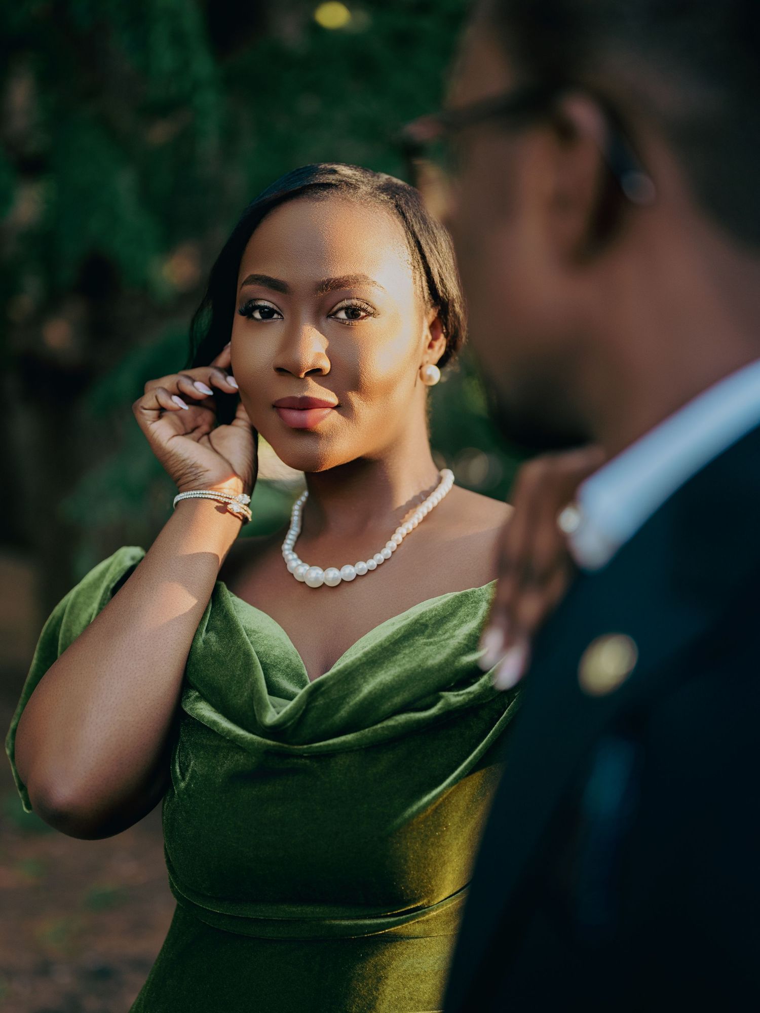 Close-up of elegant Black woman in green velvet off-shoulder dress wearing pearl necklace in golden outdoor light.