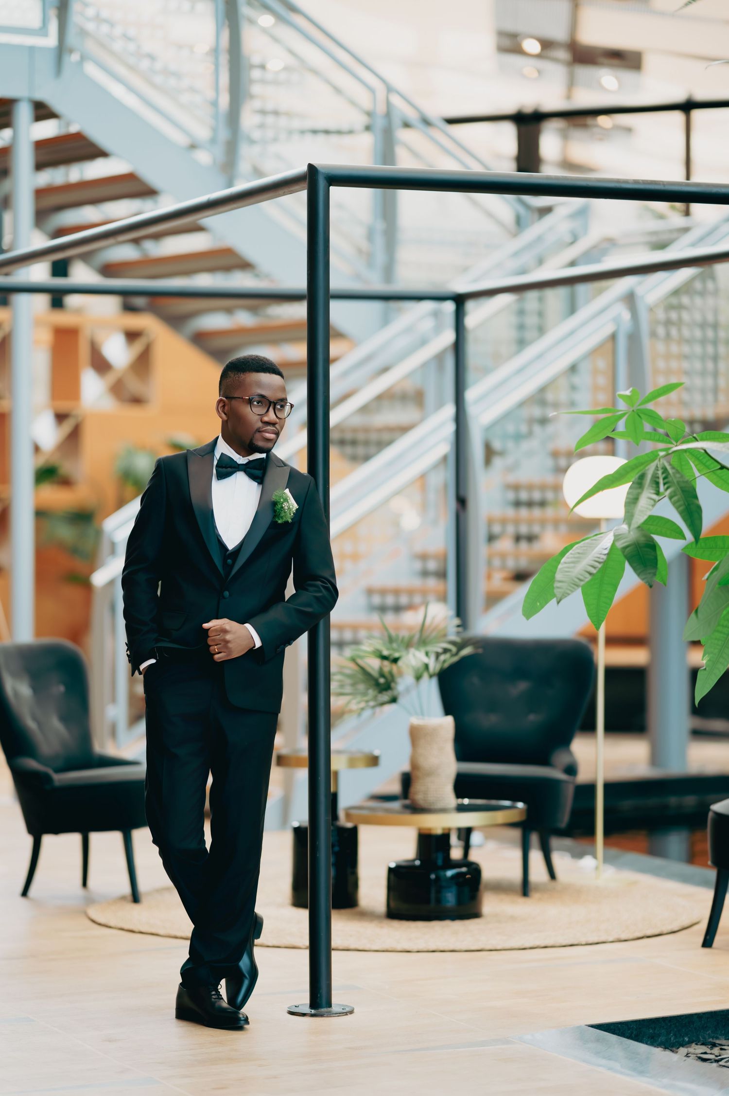 Stylish groom in black tuxedo with bow tie and boutonniere posing in a modern hotel lobby.