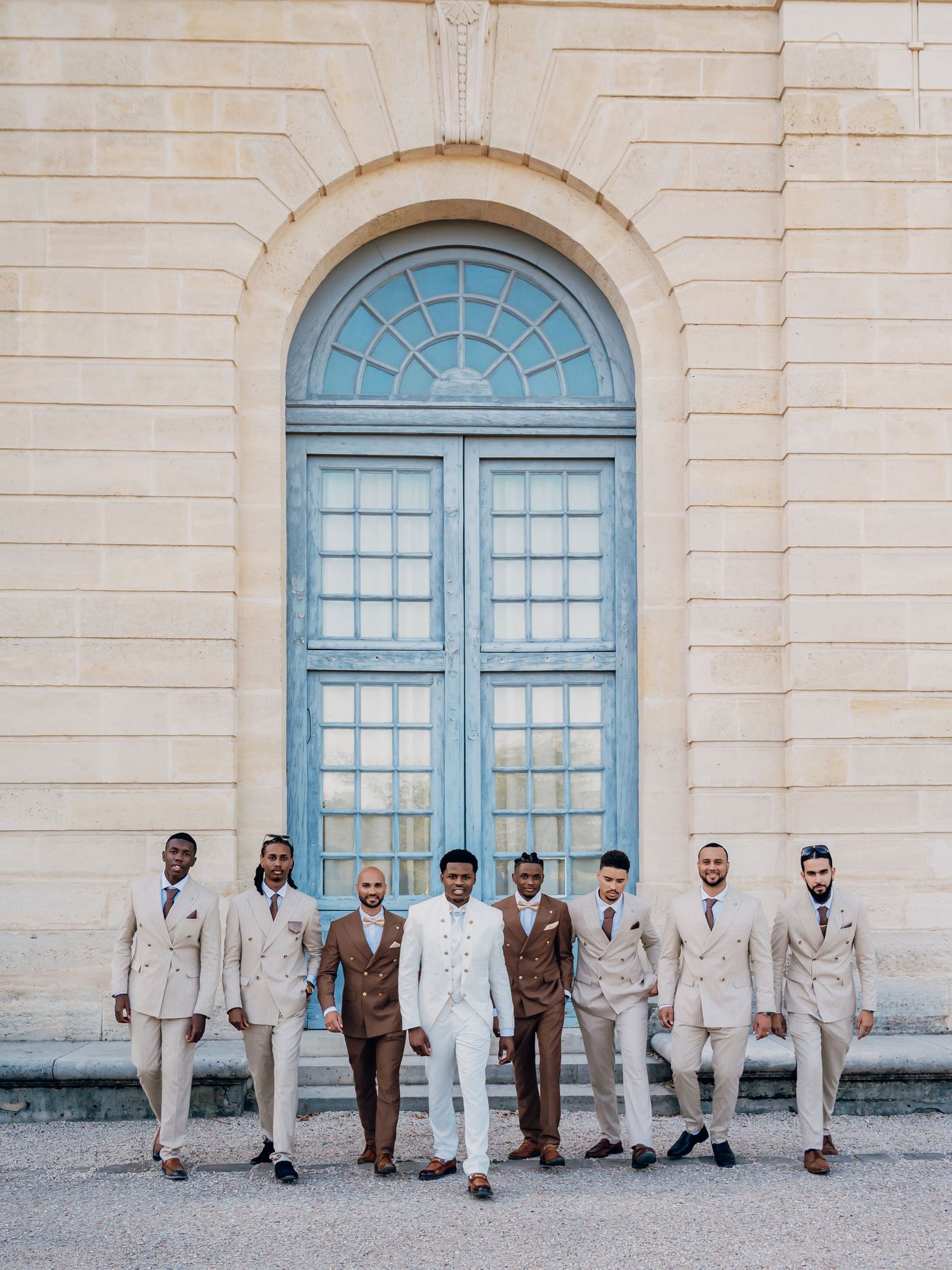 Groom in white suit leads groomsmen in tan and brown suits before an arched blue door.