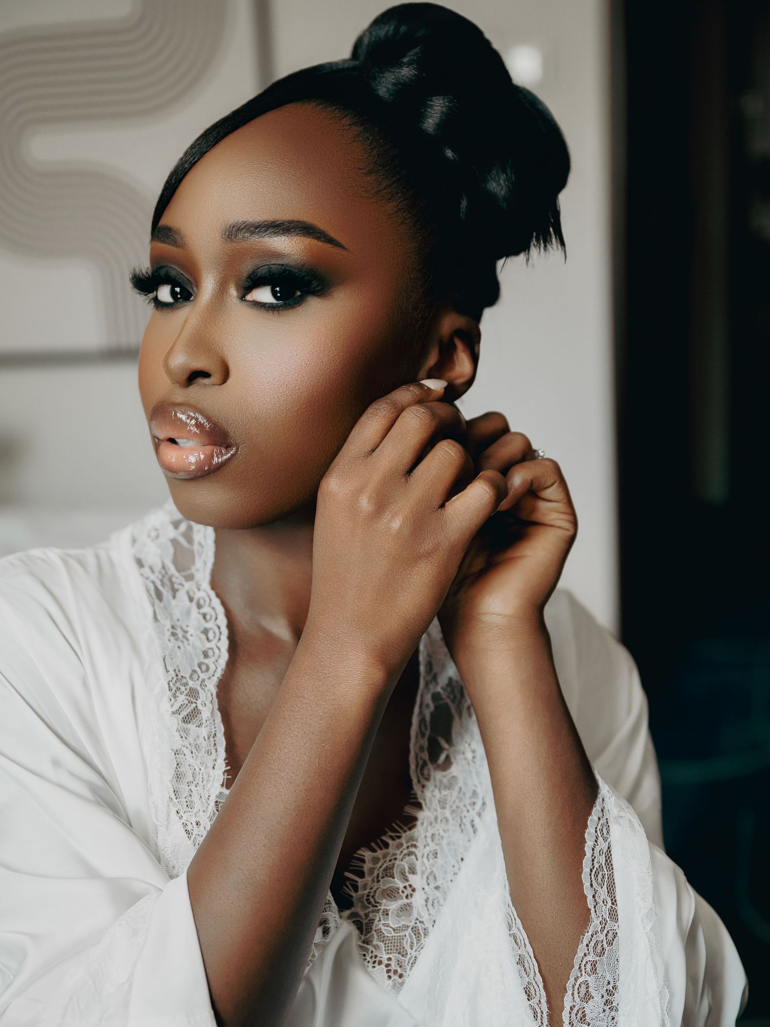 Bride in white lace robe putting on earrings before wedding.