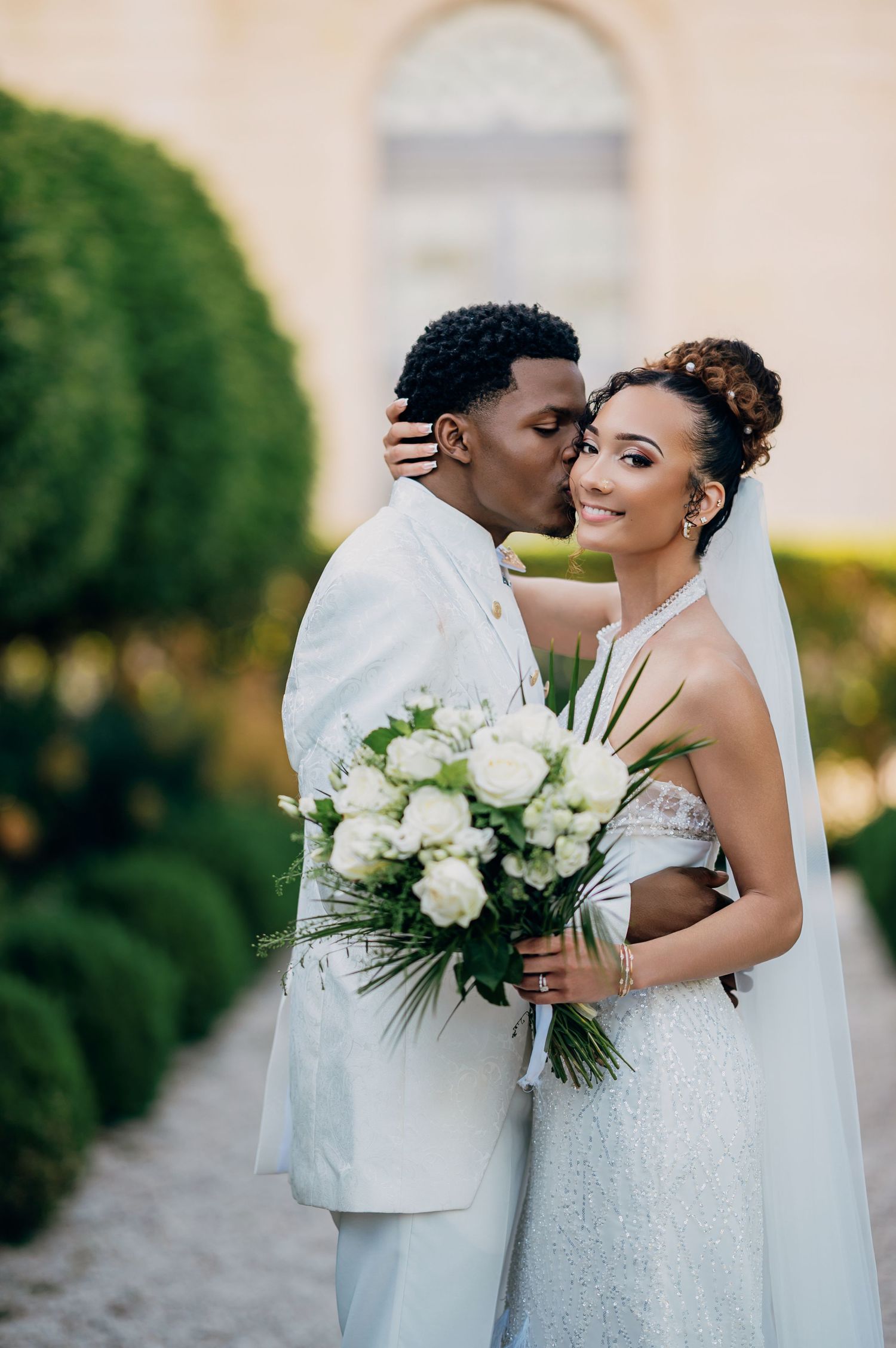 Groom in white suit kisses bride's cheek as she smiles holding white rose bouquet in garden setting.