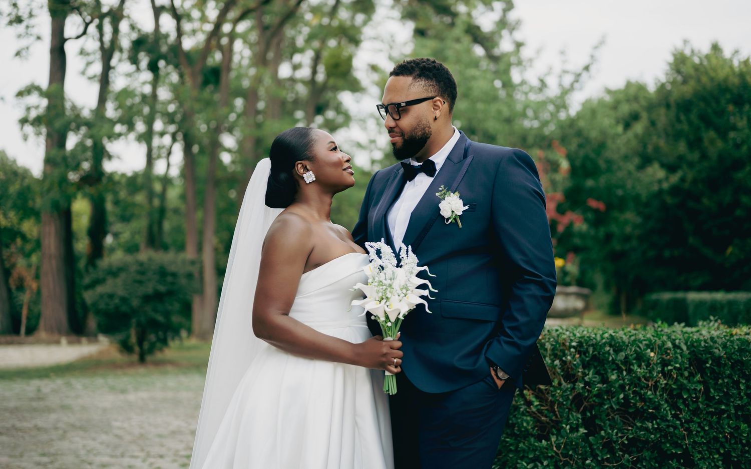 A Black bride and groom share a romantic moment outdoors, she in a white gown, he in a navy suit.