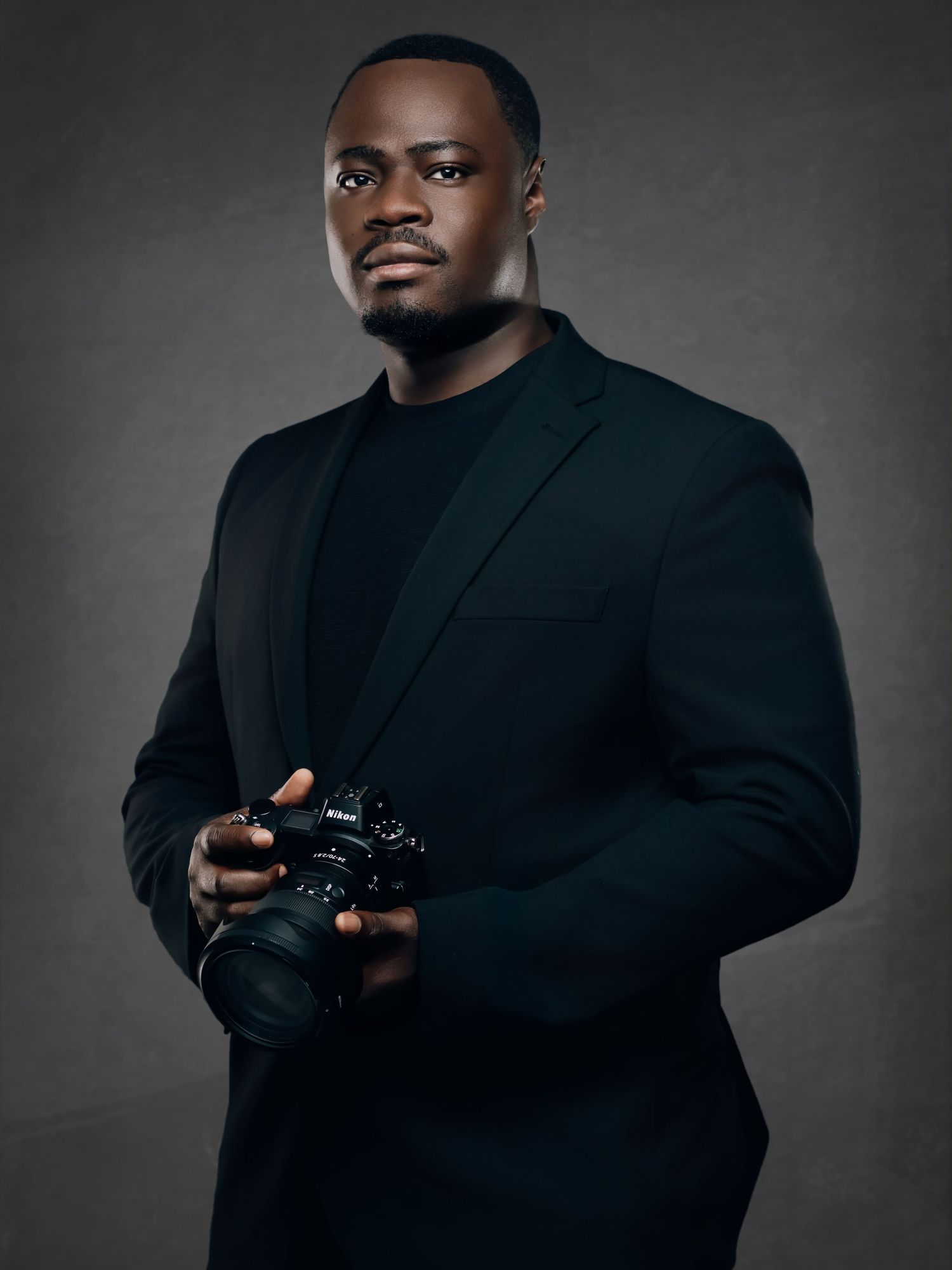 A Black male photographer in a black blazer holds a Nikon camera in a studio setting.