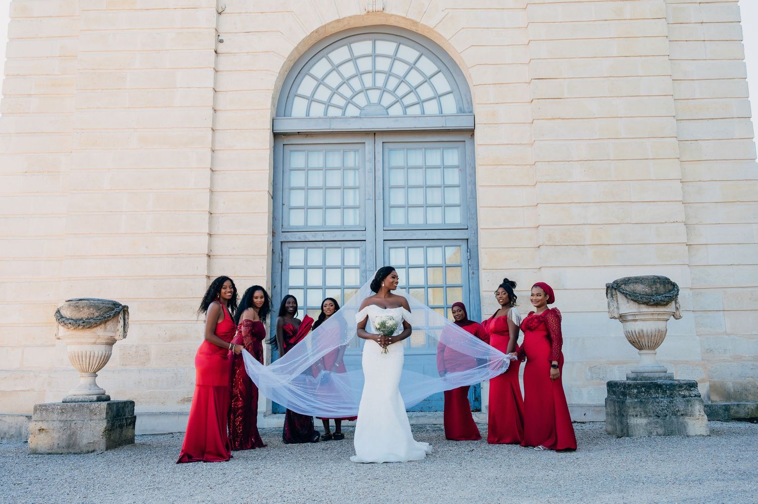 Bride in white gown with bridesmaids in red dresses spreading veil before ornate doors.