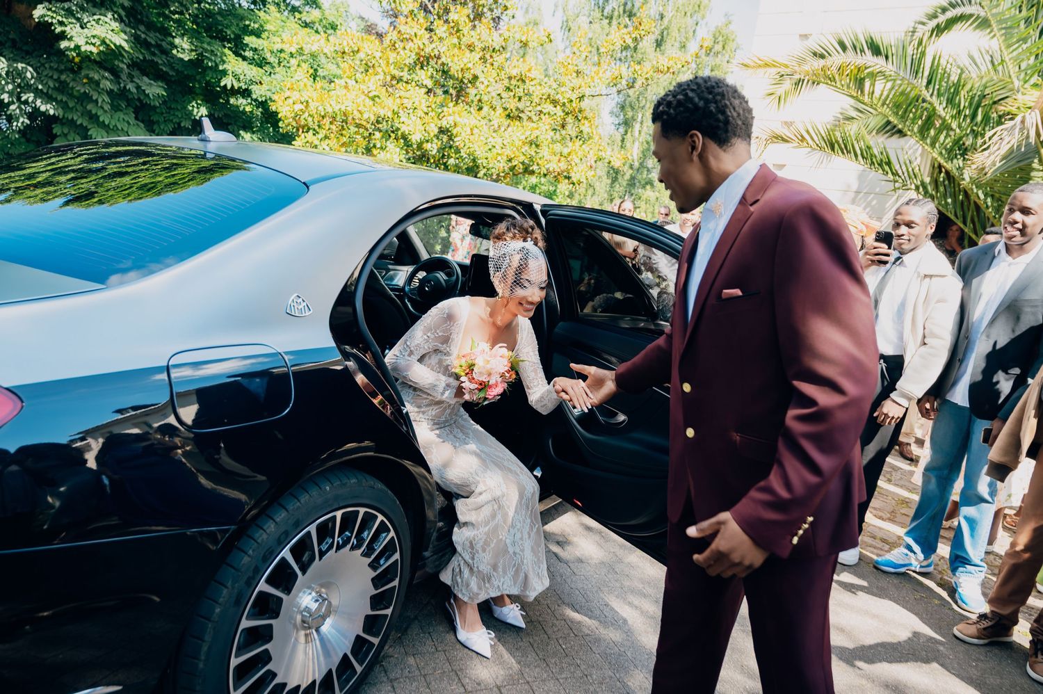 Groom in burgundy suit helping bride exit luxury black wedding car outdoors.