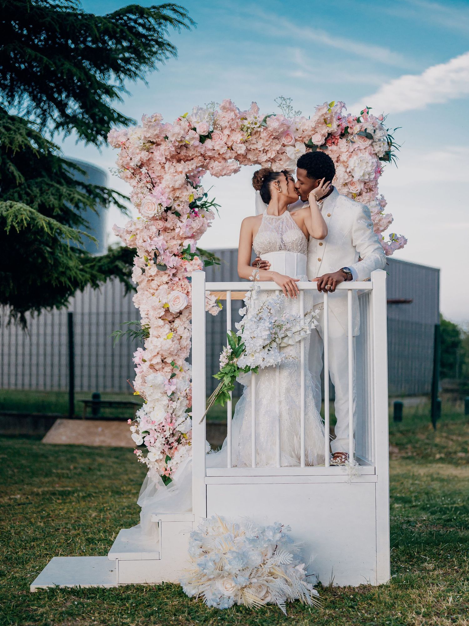 Couple kissing under a floral arch with pink blooms at an outdoor wedding ceremony.