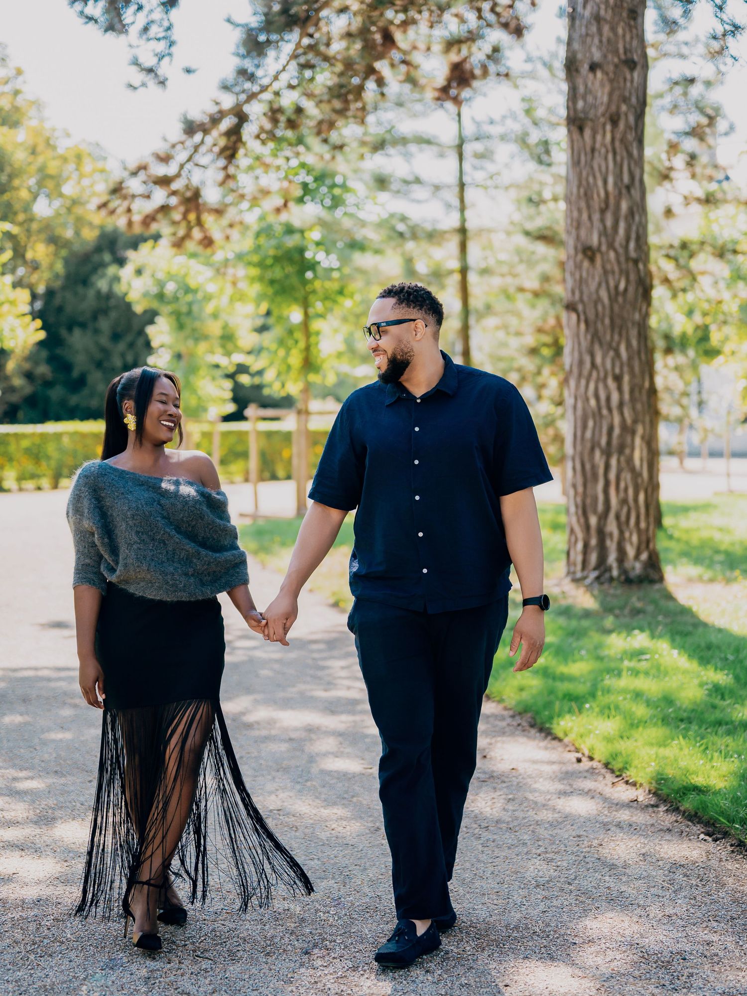 Laughing Black couple strolling hand in hand along a tree-lined path, woman in grey wrap and black fringe skirt.