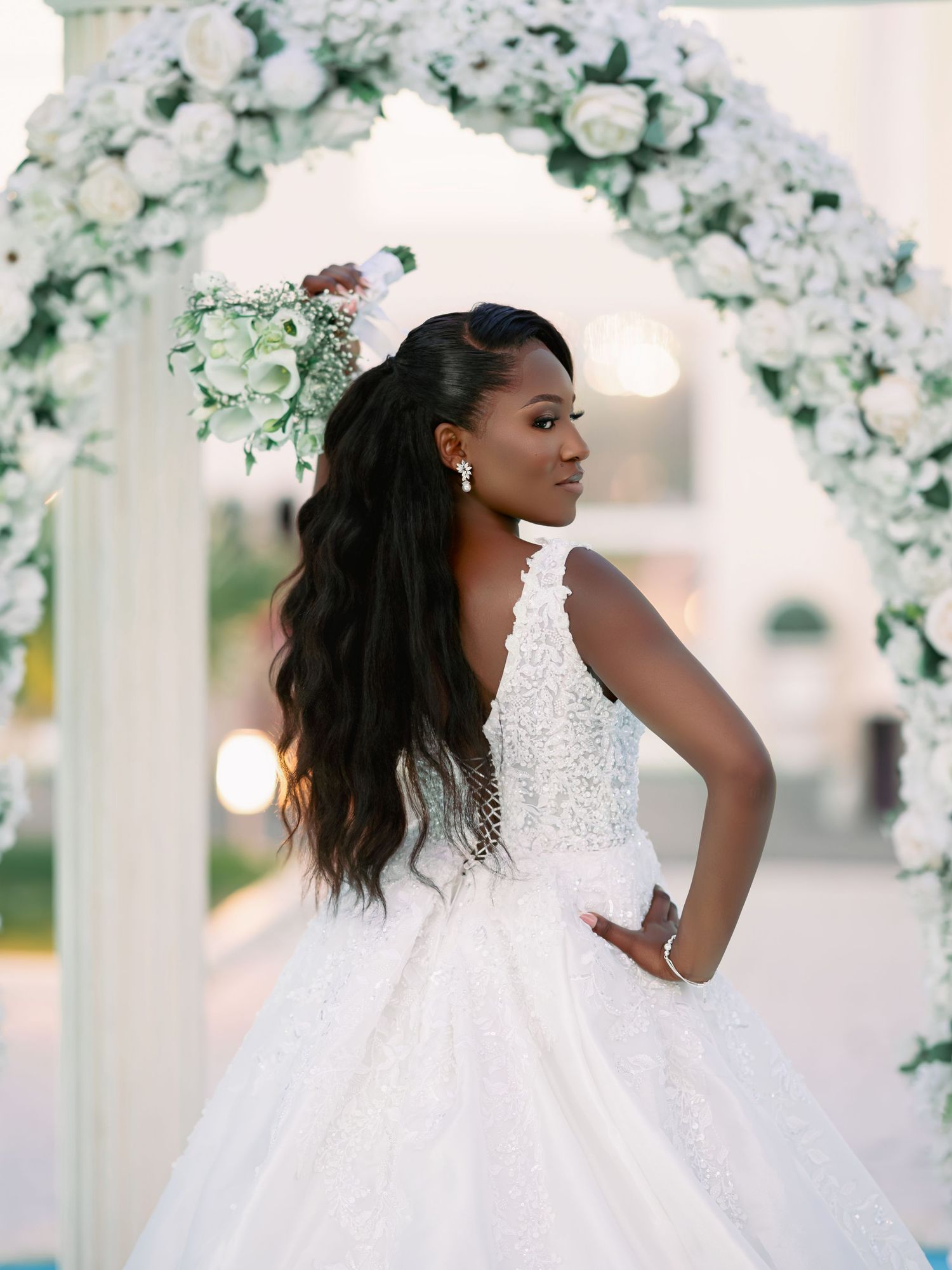 Bride in beaded ballgown poses under white floral arch with long flowing hair.