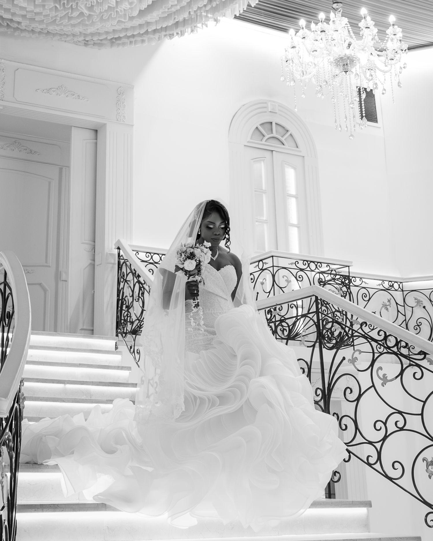 Black and white photo of Black bride in ruffled gown descending grand staircase with chandelier above.