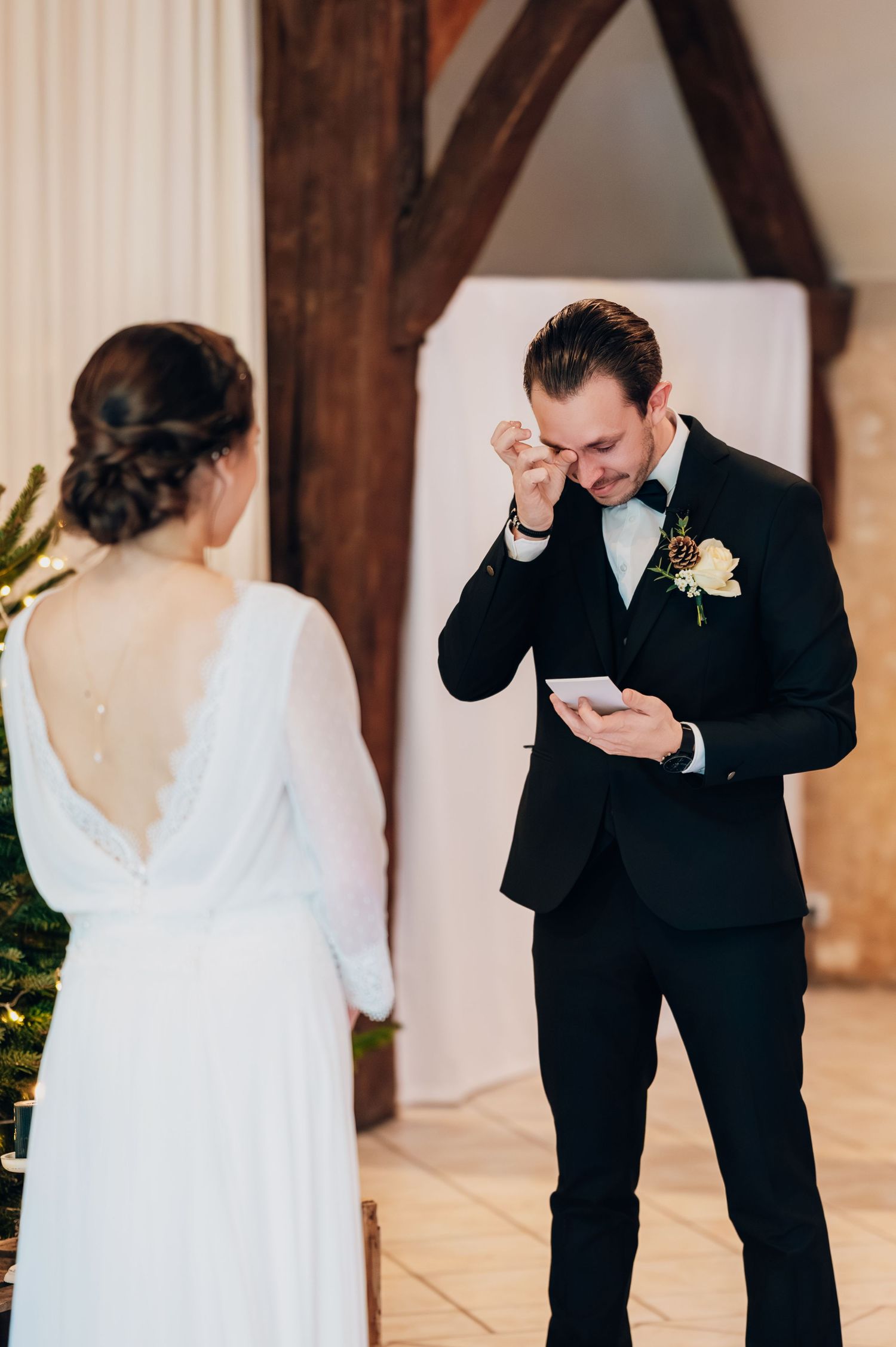 Emotional groom wiping tears while reading wedding vows to bride in elegant backless gown.