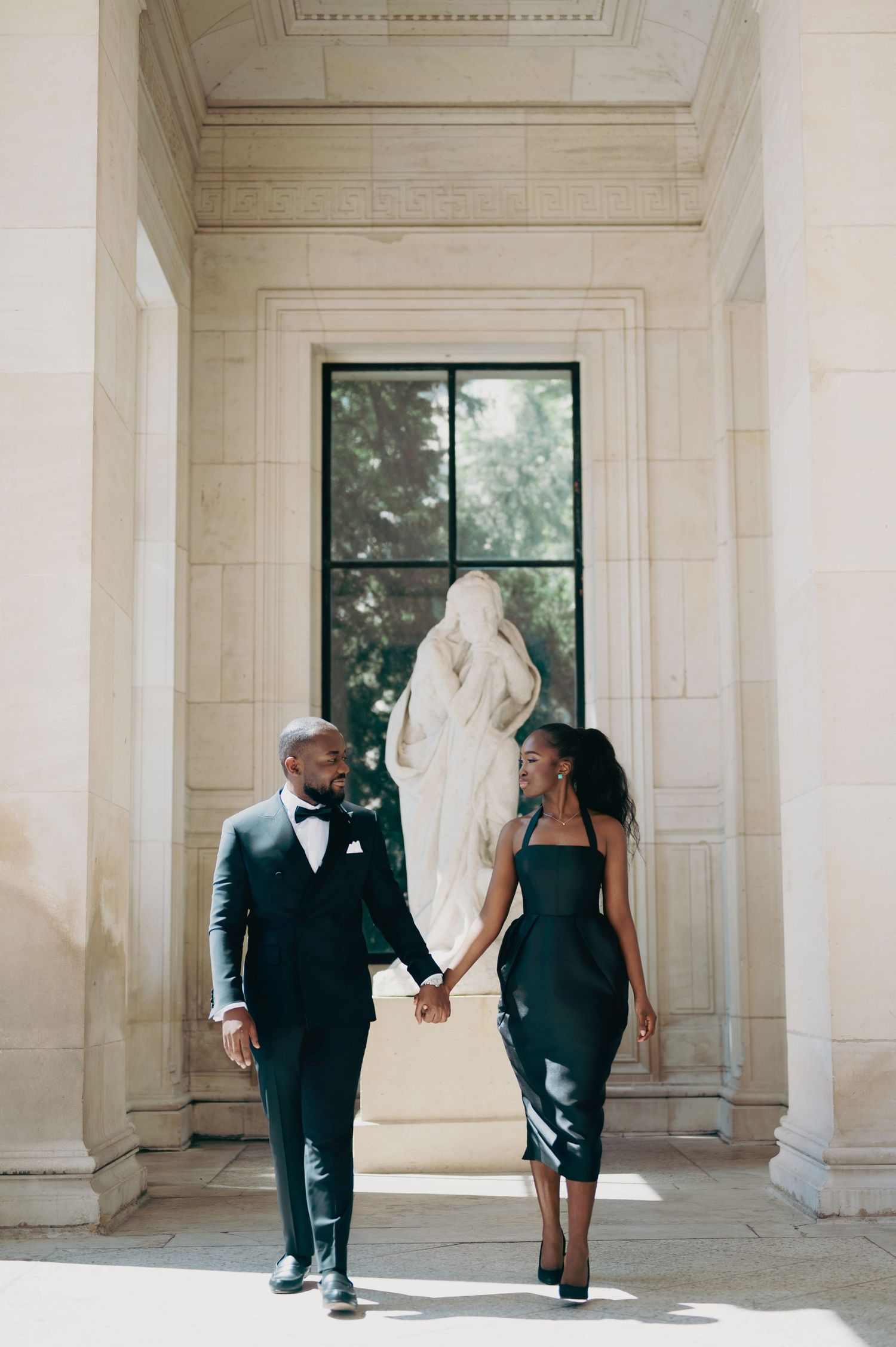 Elegant couple in matching dark green formal attire holding hands while walking past a classical marble statue indoors.