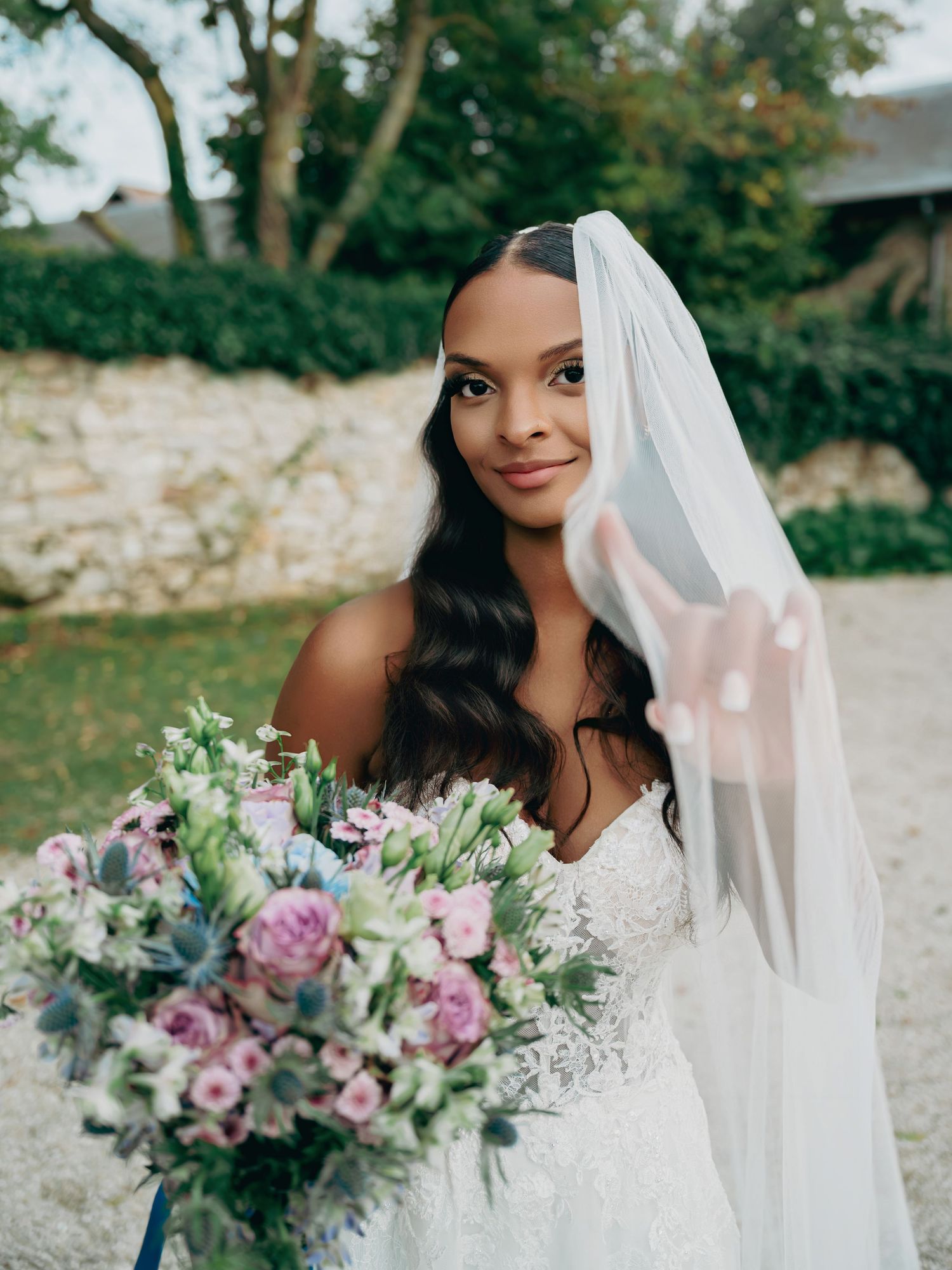 Radiant Black bride in off-shoulder lace gown holding purple floral bouquet with flowing veil in garden setting.