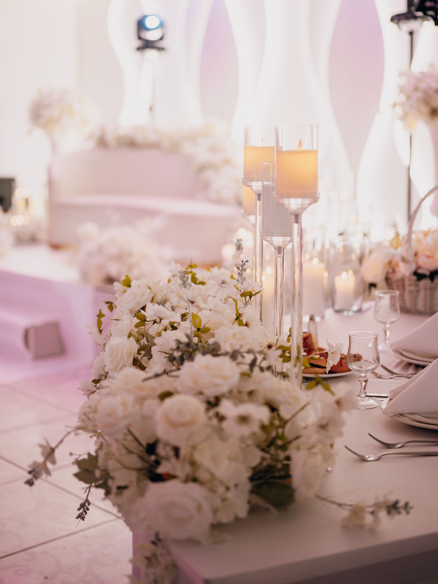 Wedding reception table with white floral centerpiece and tall glass candle holders in an all-white elegant venue.