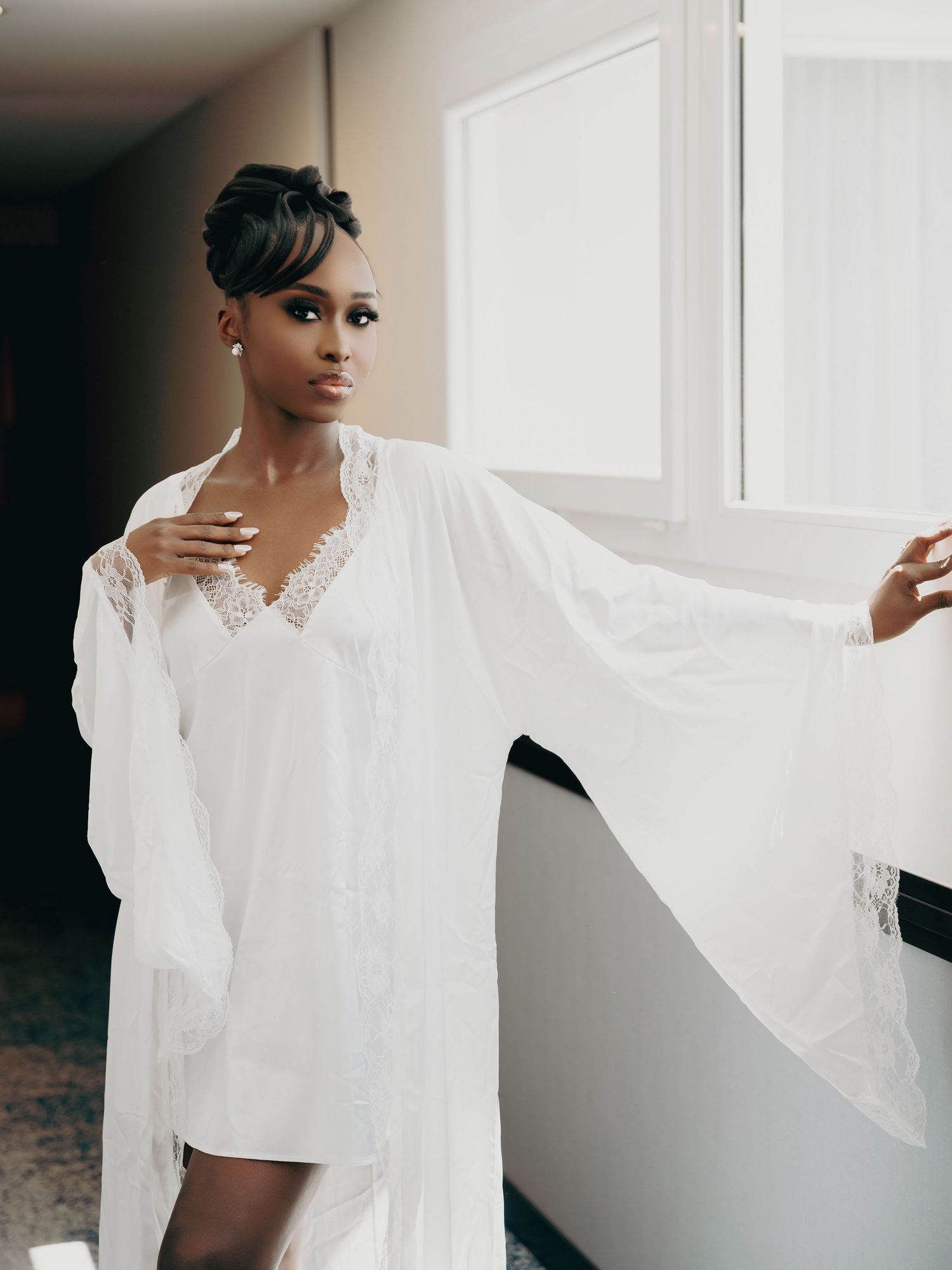 Elegant Black bride poses in flowing white lace robe by a sunlit window before wedding.