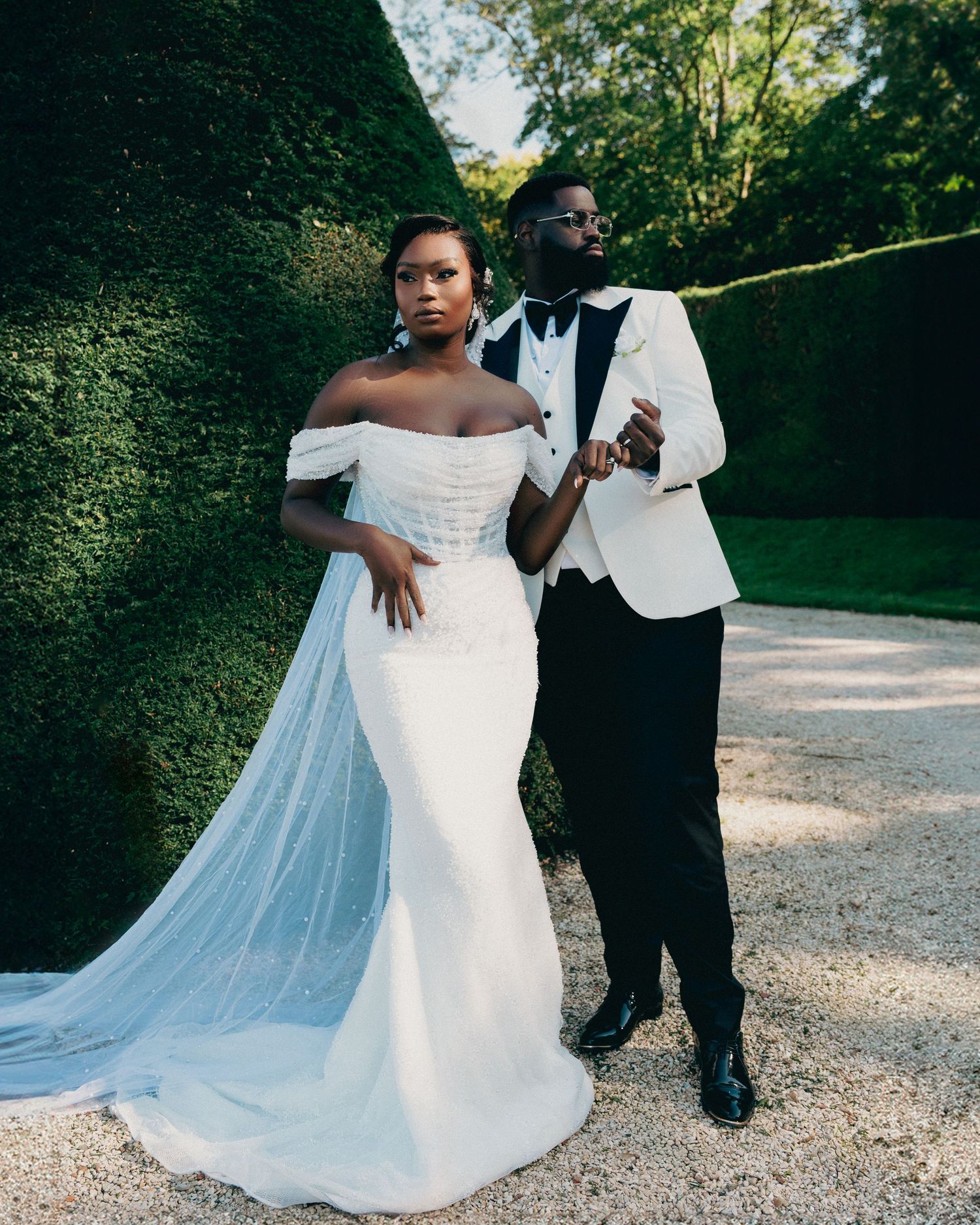 Bride in mermaid gown with flowing veil and groom in white tuxedo posing by tall hedges.