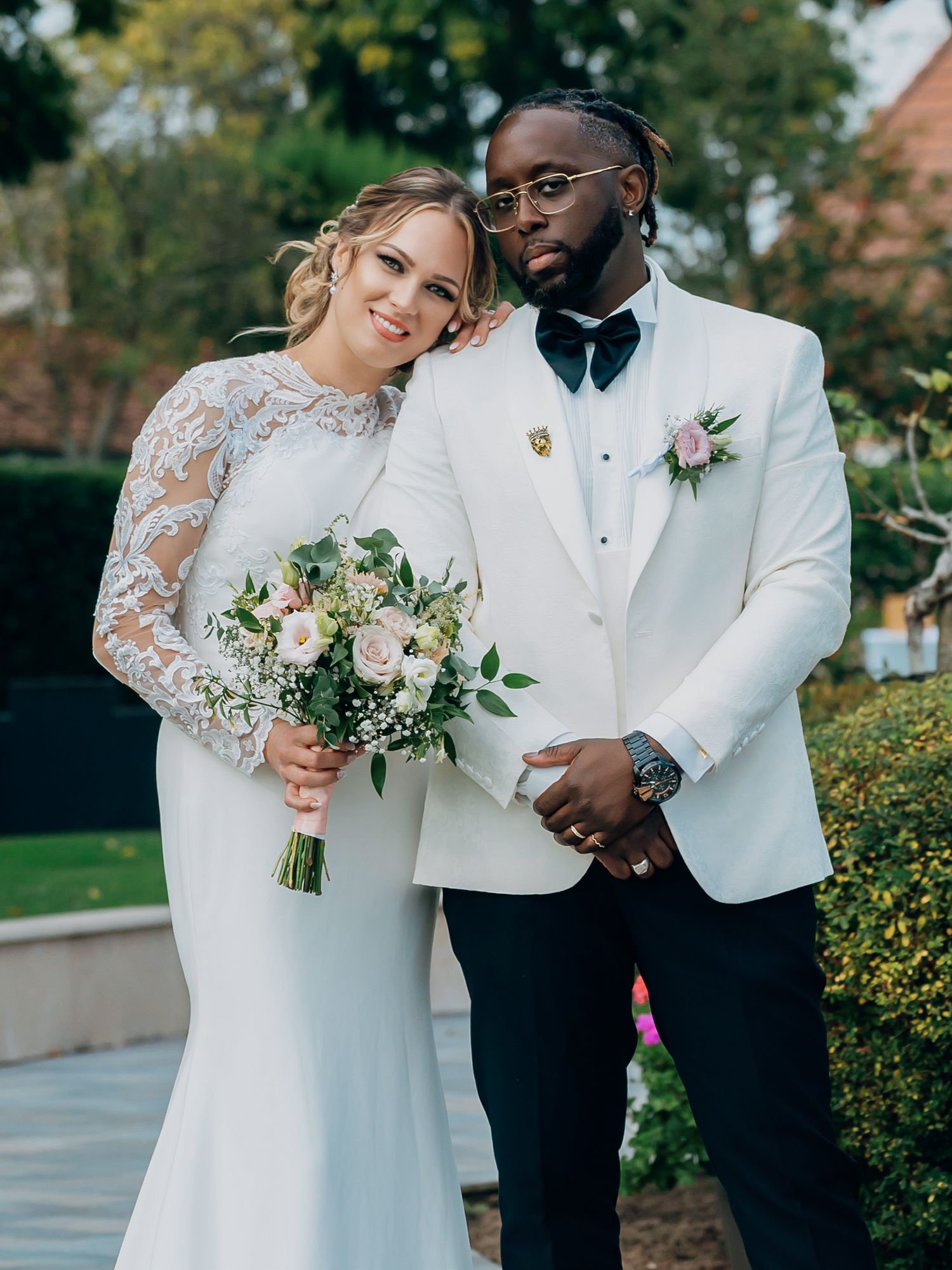 Interracial wedding couple posing outdoors, bride in lace long-sleeve gown holding pink bouquet, groom in white tuxedo.