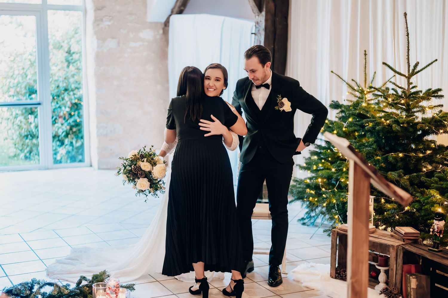 Bride and groom sharing warm embrace with guest near Christmas tree at winter wedding celebration.