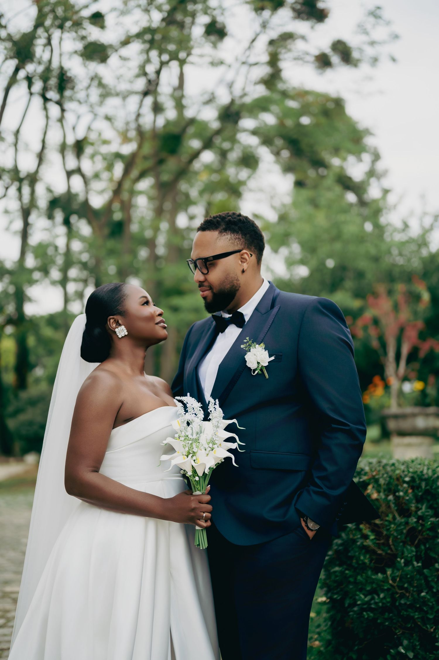 Black bride and groom sharing a loving gaze outdoors, bride holding white calla lily bouquet in elegant strapless gown.
