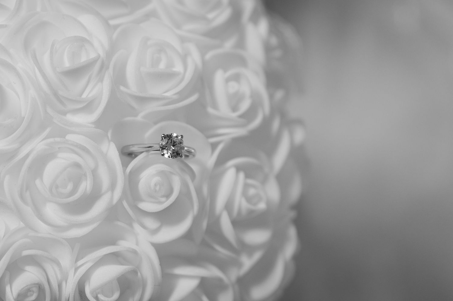 Black and white close-up of diamond solitaire engagement ring resting on white rose bouquet.