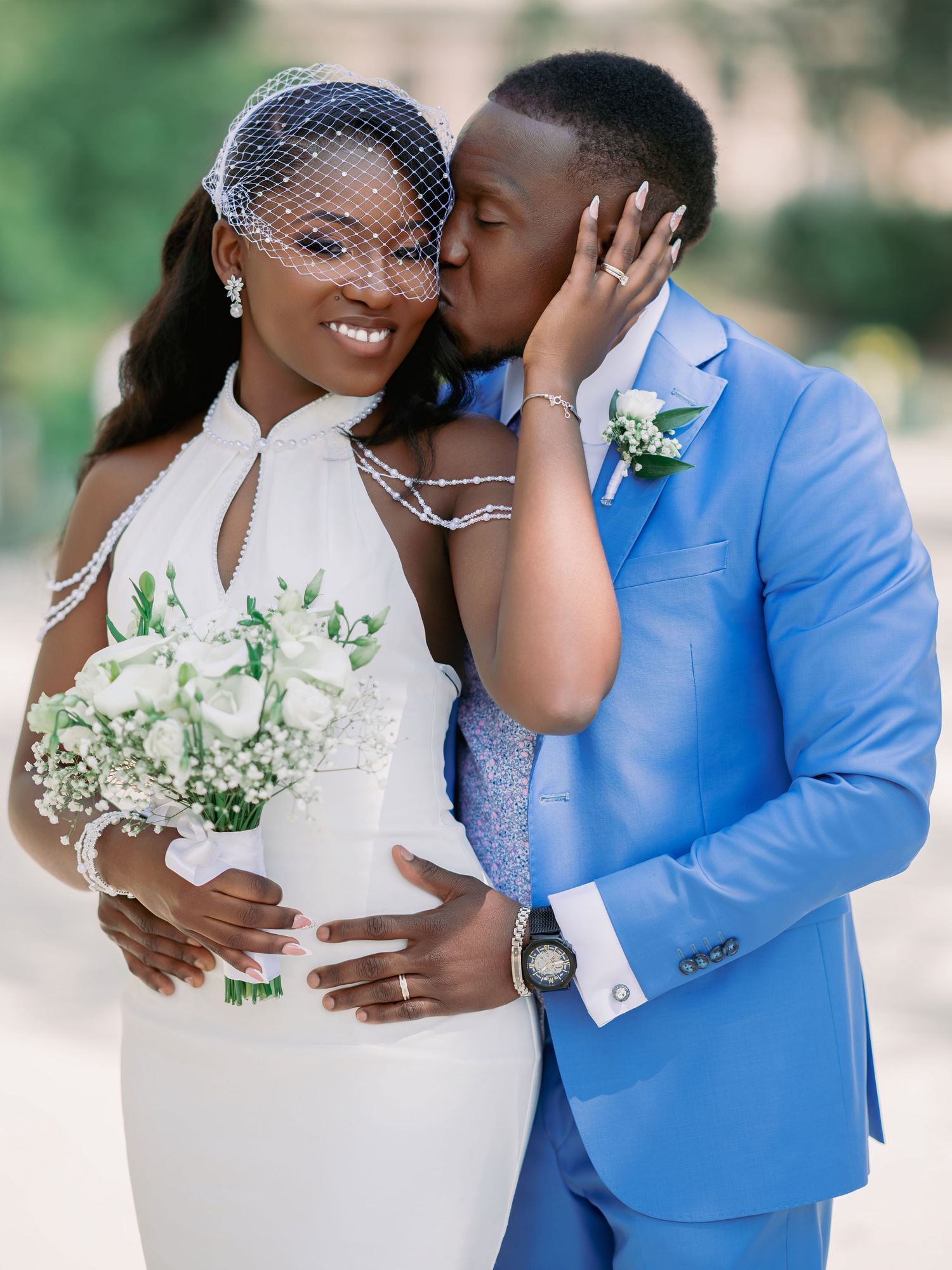 Groom in blue suit kissing bride wearing birdcage veil and white gown with bouquet.