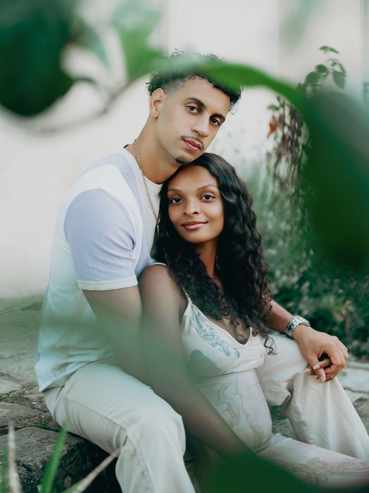 Young couple sitting close together surrounded by lush green foliage in a romantic outdoor setting.