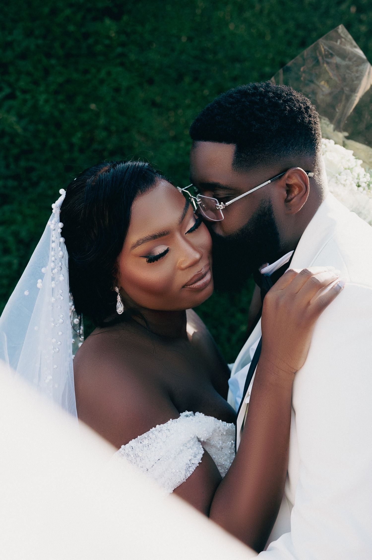 Intimate moment between Black newlyweds under a veil, groom in glasses kisses bride's cheek.