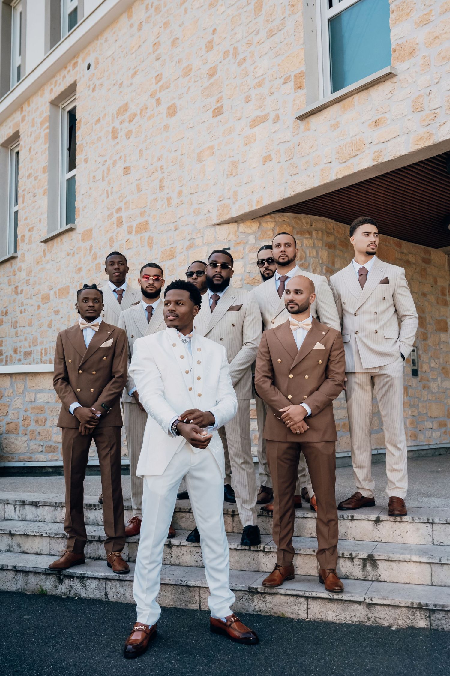Groom in white suit standing confidently before groomsmen in tan and brown double-breasted suits on stone steps.