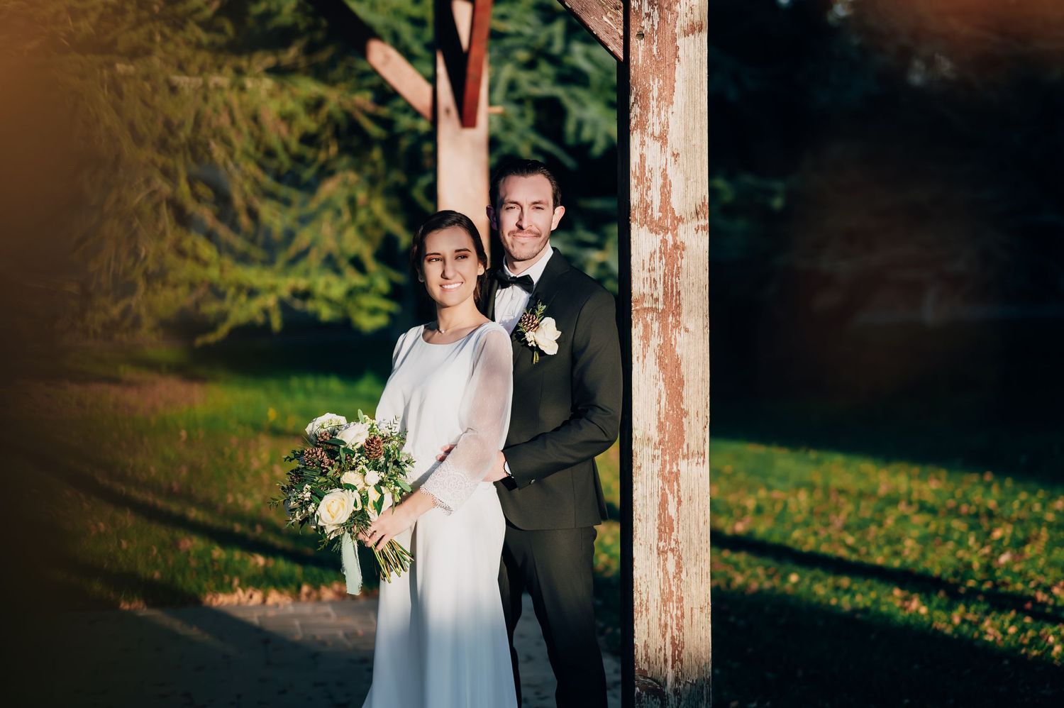 Elegant wedding couple standing between wooden posts in a lush green park, bride holding bouquet.