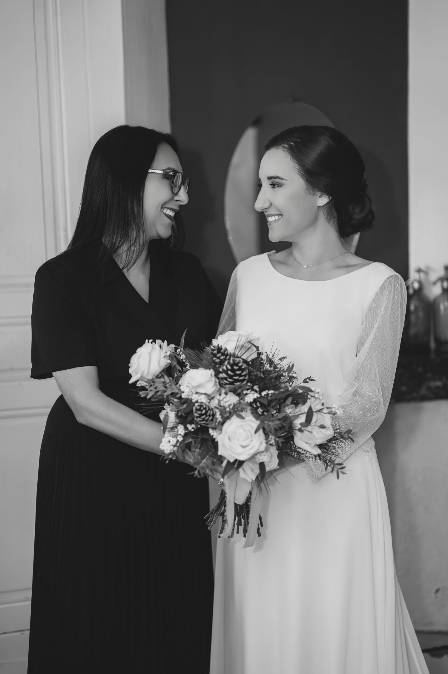 Black and white photo of bride in white gown and woman in black dress laughing together holding winter bouquet.