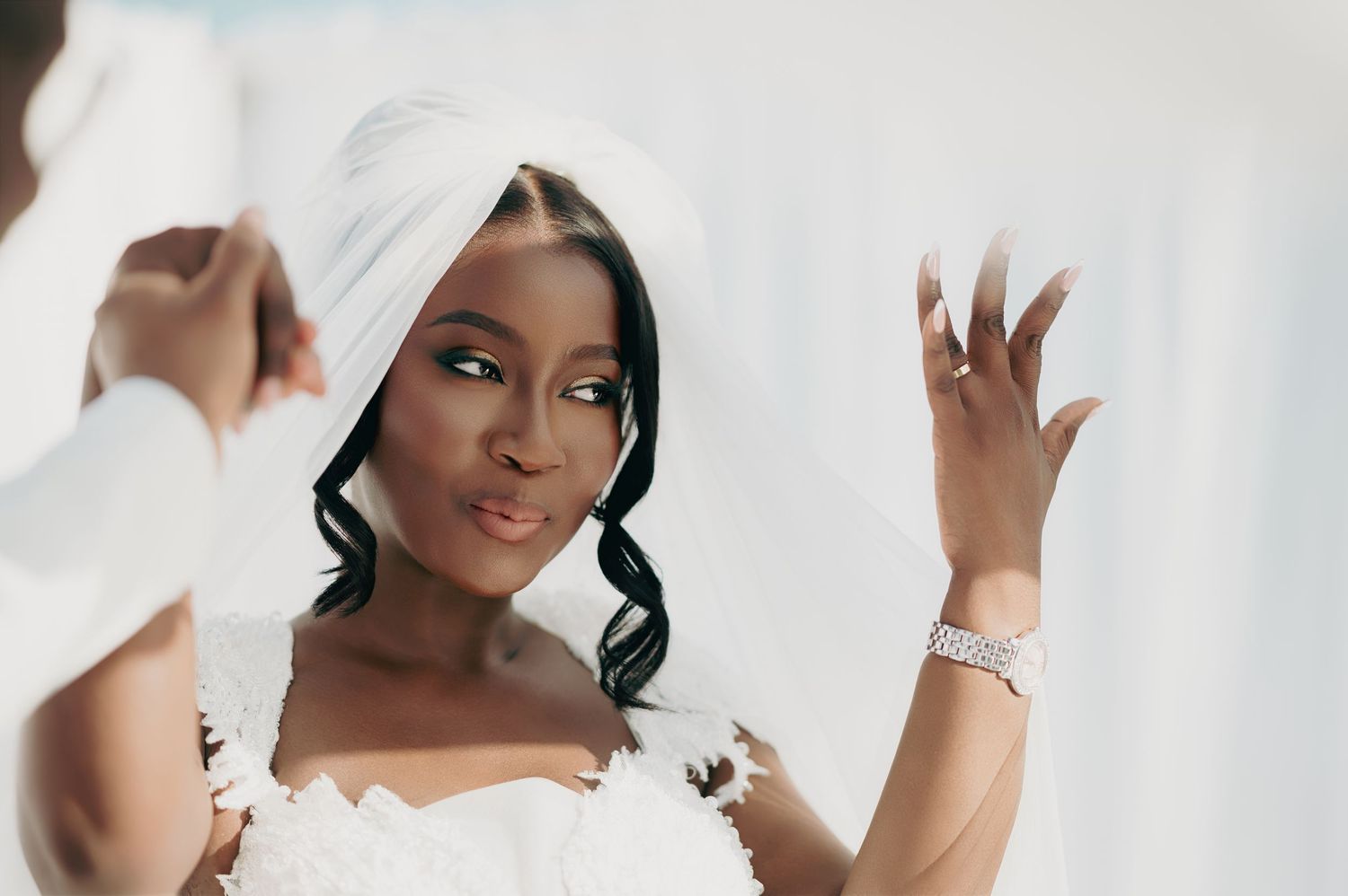 A bride in white with a veil wrapped around her head applies makeup while admiring herself in a mirror.