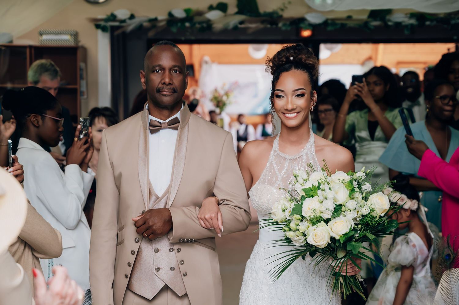 Bride in white halter gown holding bouquet walks aisle with escort in tan suit.