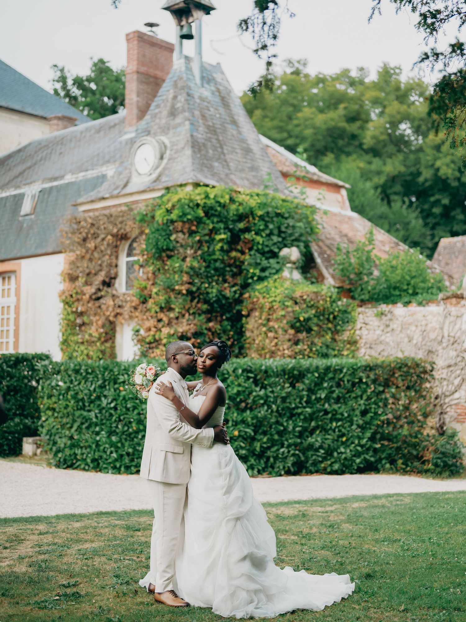 Bride and groom share a romantic kiss on lush green lawn before ivy-covered French chateau wedding venue.