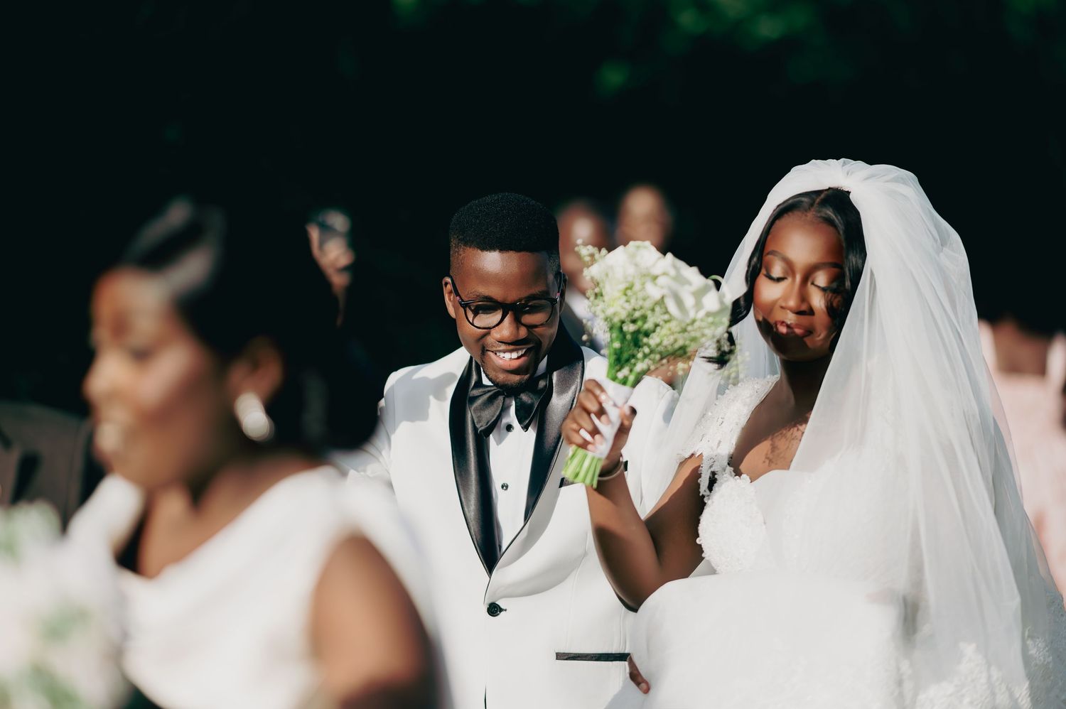 A joyful groom in a white tuxedo walks beside his bride in a white veil during their wedding ceremony.