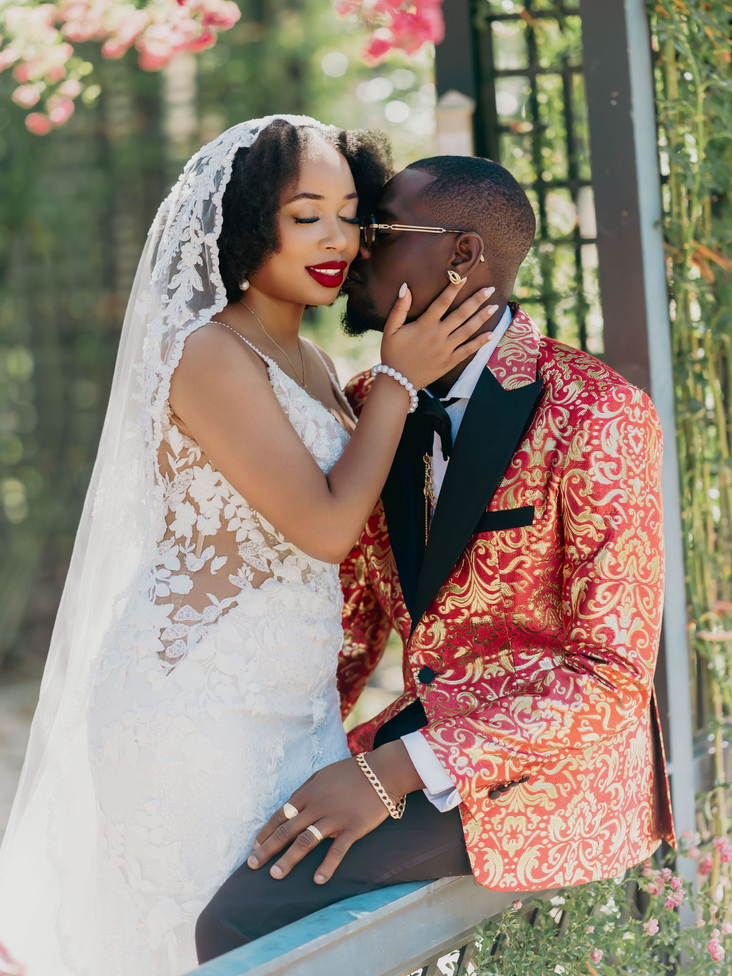 Bride in lace gown and veil shares romantic kiss with groom in ornate red gold blazer at outdoor wedding.