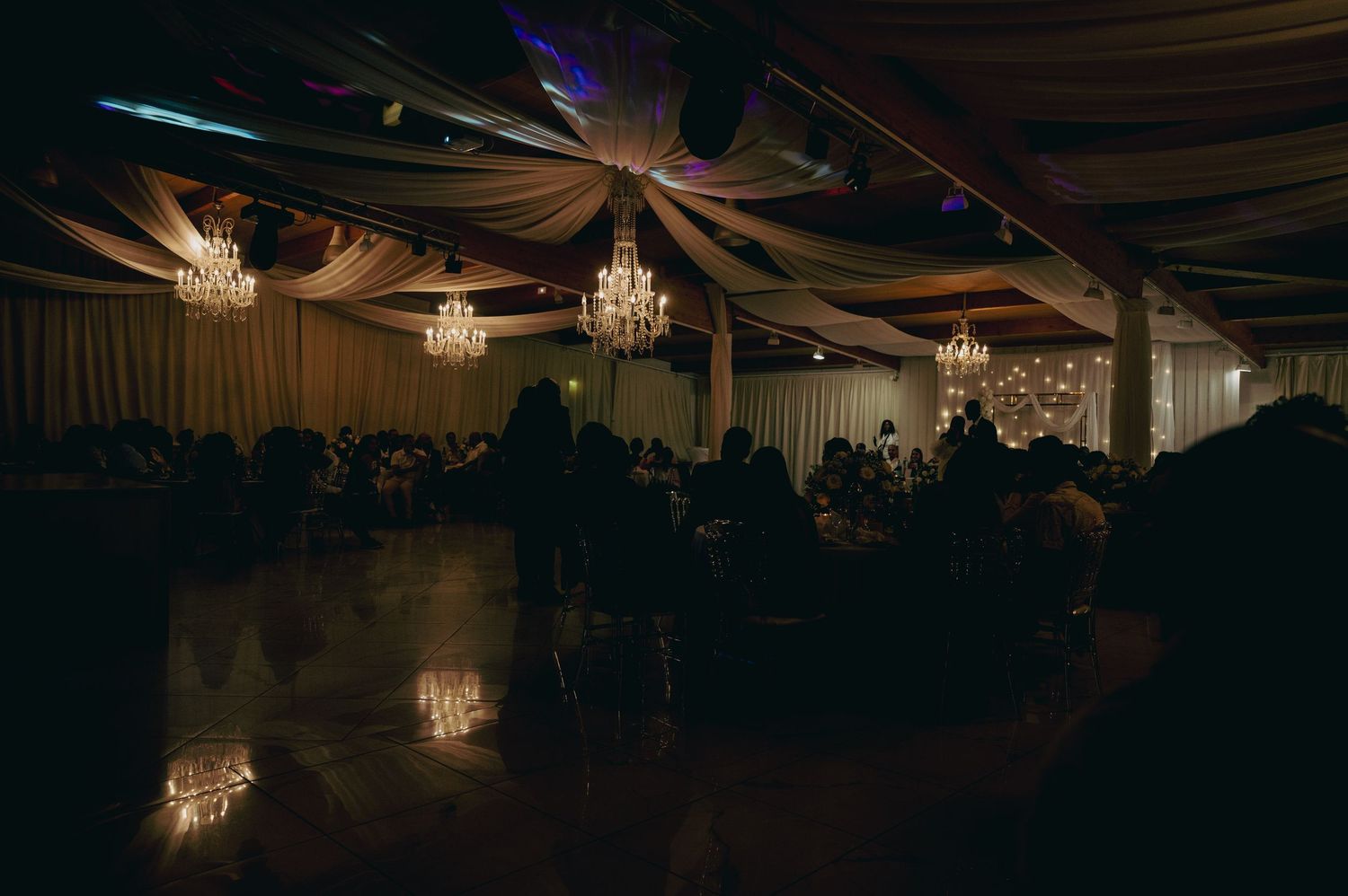 Elegant wedding reception hall with crystal chandeliers and draped ceiling at night.