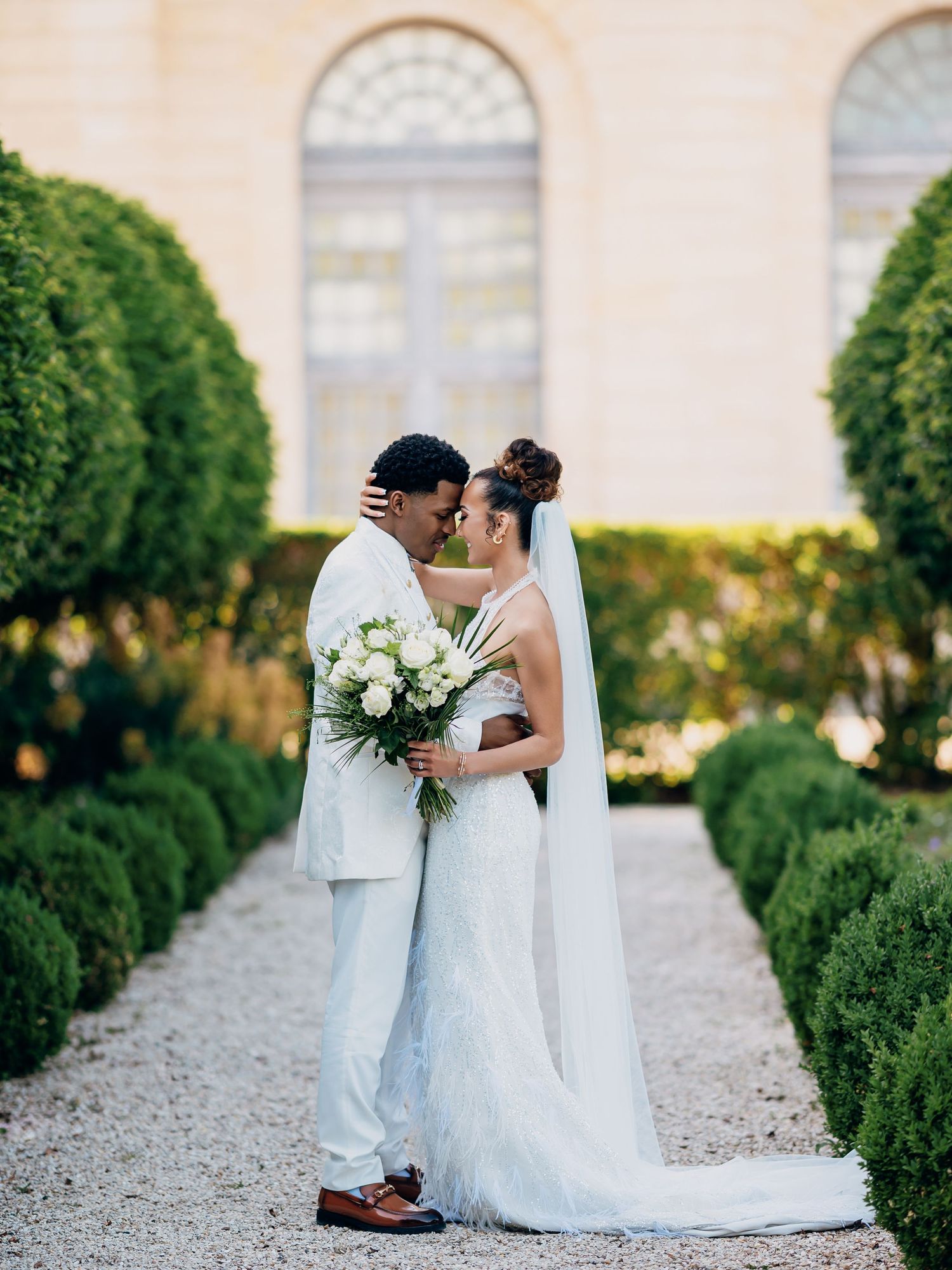 Bride and groom share a kiss holding white rose bouquet in formal garden pathway.