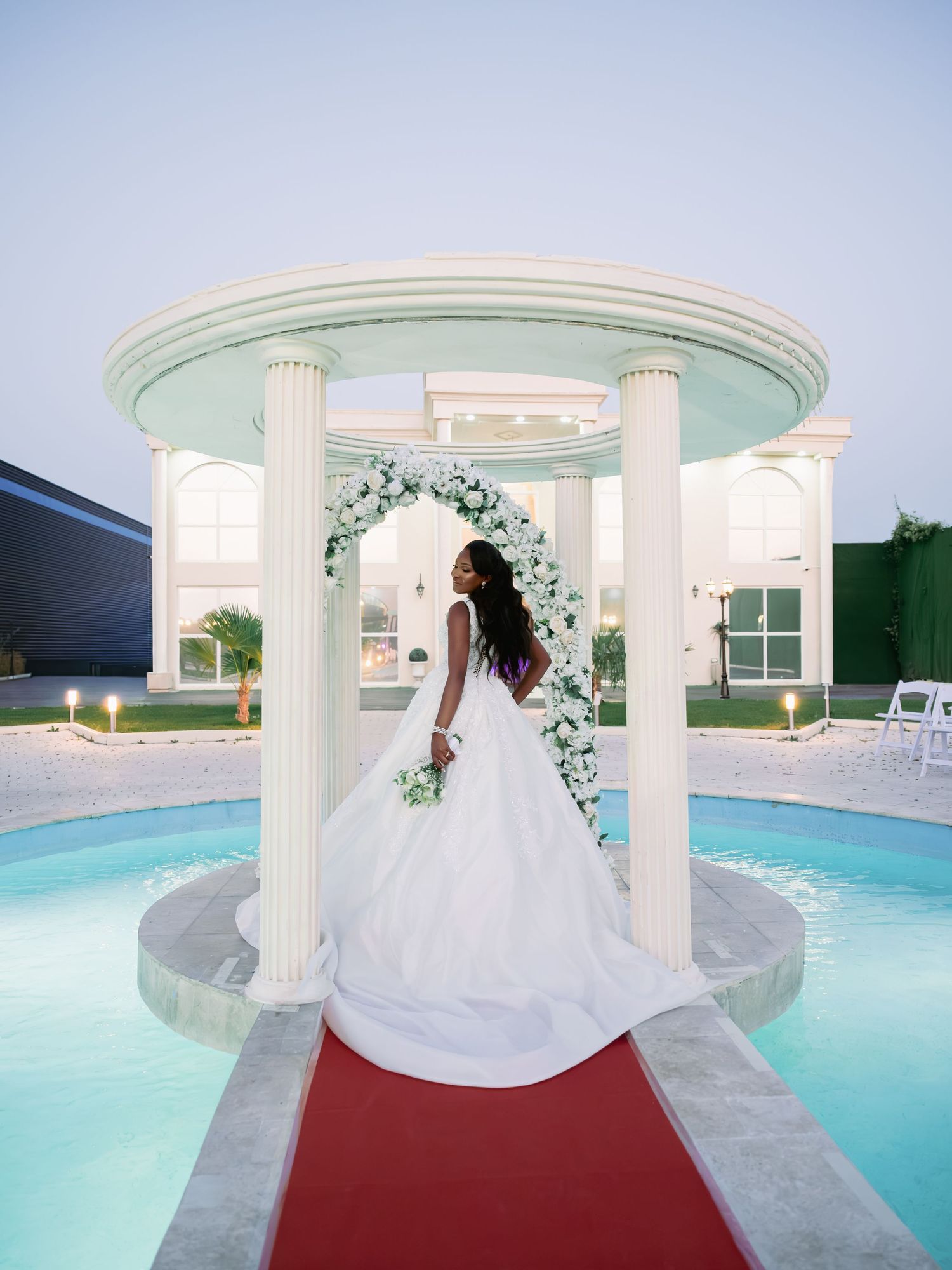 Bride in white ball gown stands under a floral arch gazebo beside a pool at twilight wedding venue.