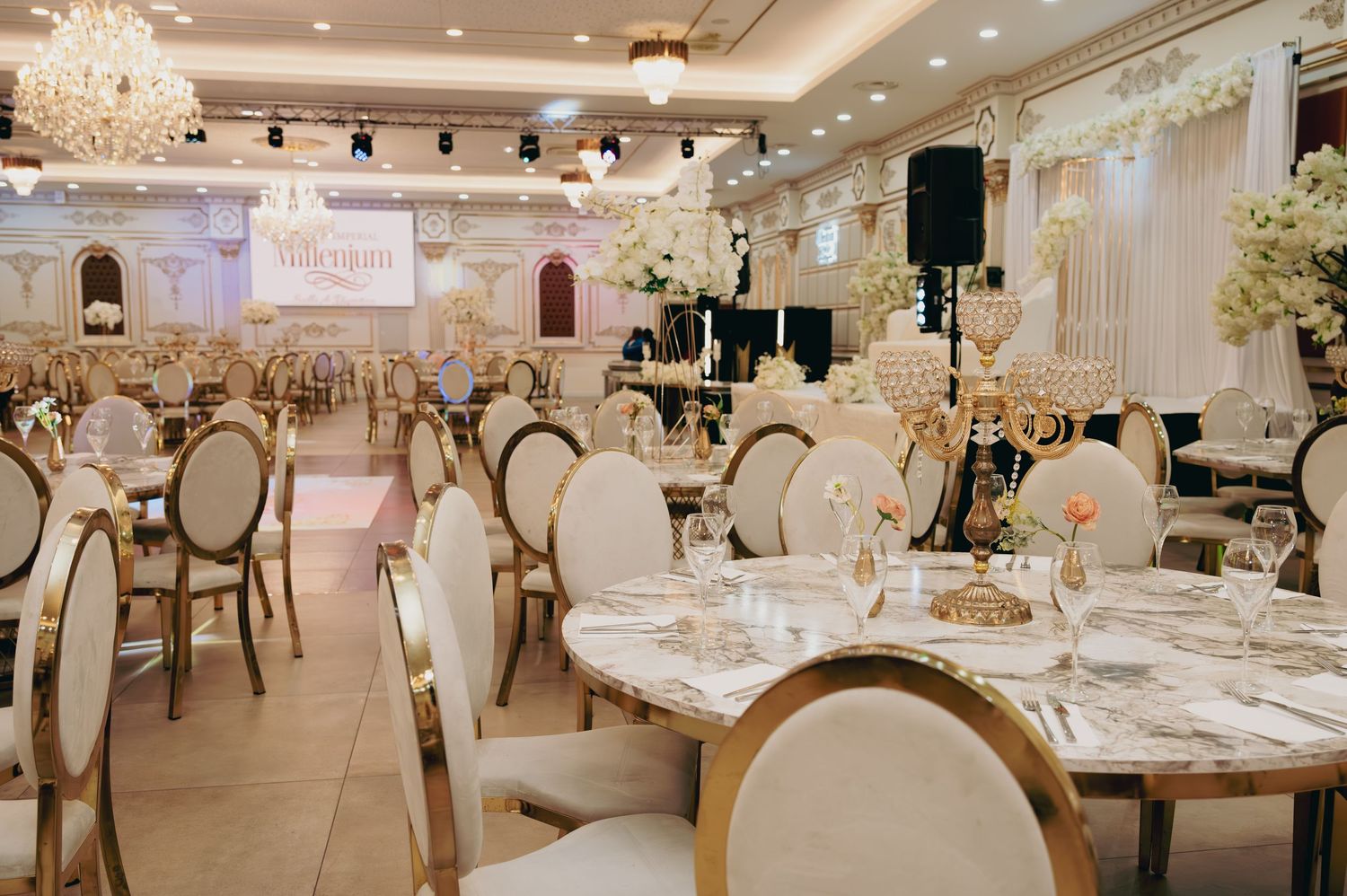 Grand wedding reception hall with marble tables, gold candelabras, and white floral centerpieces.