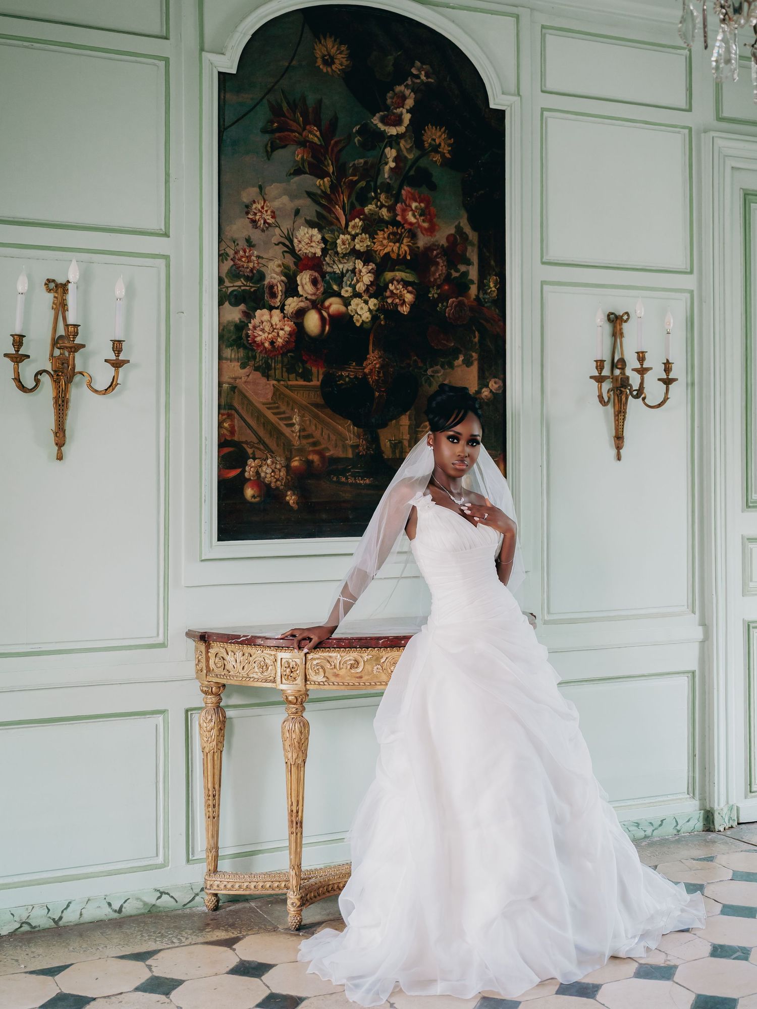 Elegant bride in white ball gown with veil posing in mint green French chateau interior.
