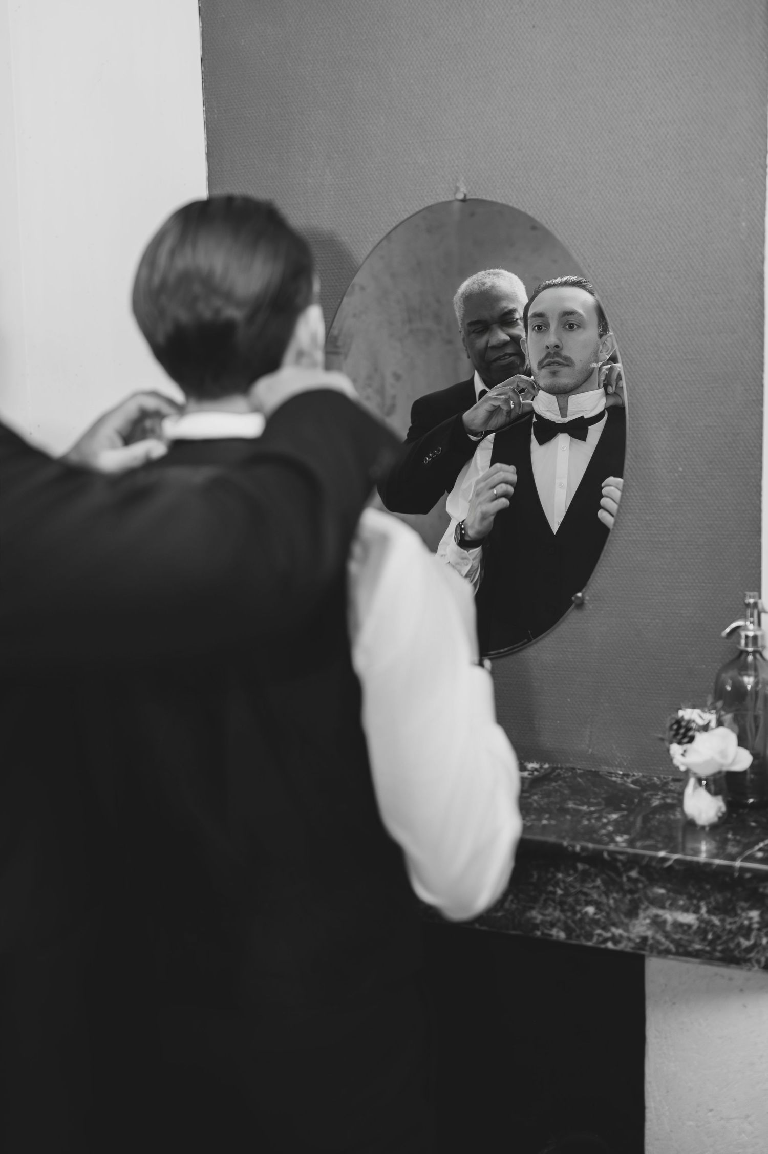 Black and white photo of groom getting bow tie adjusted by older man in mirror reflection.