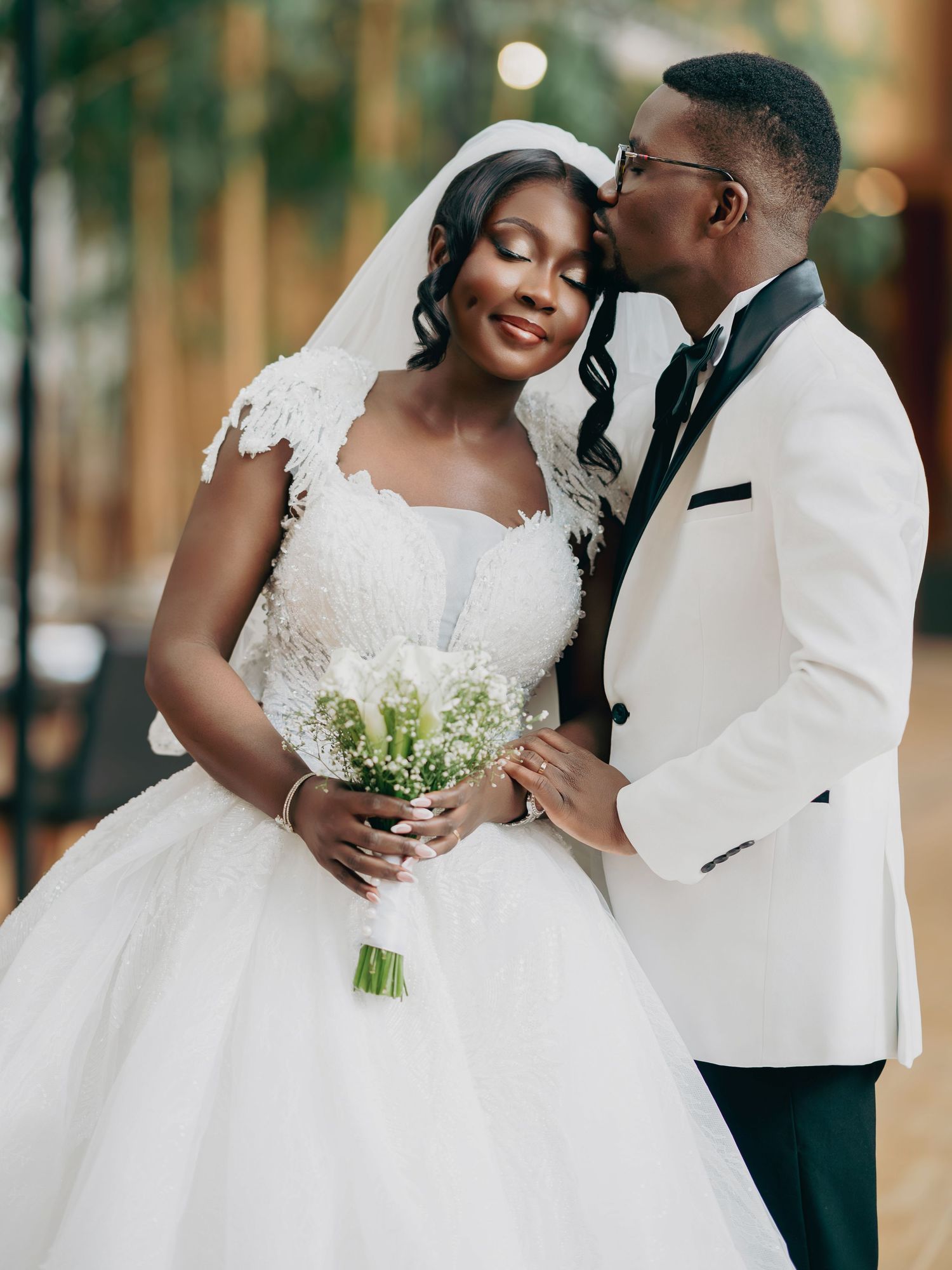 Black groom in white tuxedo kissing bride's forehead as she holds a white bouquet in romantic pose.